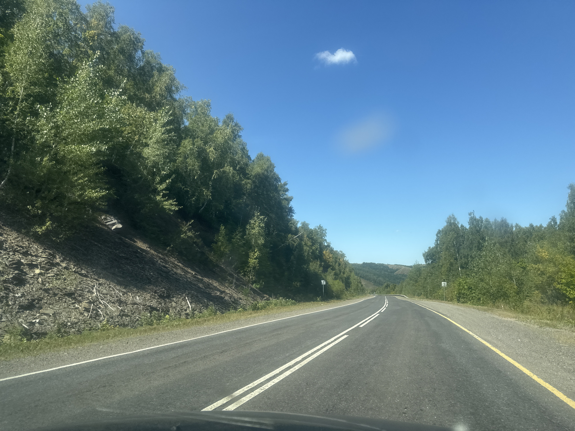 The image depicts a two-lane asphalt road curving to the right, bordered by lush green trees and a steep, rocky embankment on the left. The sky is a clear, bright blue with a single small white cloud. The road has double white lines marking the center and a yellow line on the right edge. The environment appears to be a rural or natural setting, likely outside a populated area. The view is from the perspective of someone inside a vehicle. There are no people visible in the image. The location is specified as the city of Zilair, Russia.
