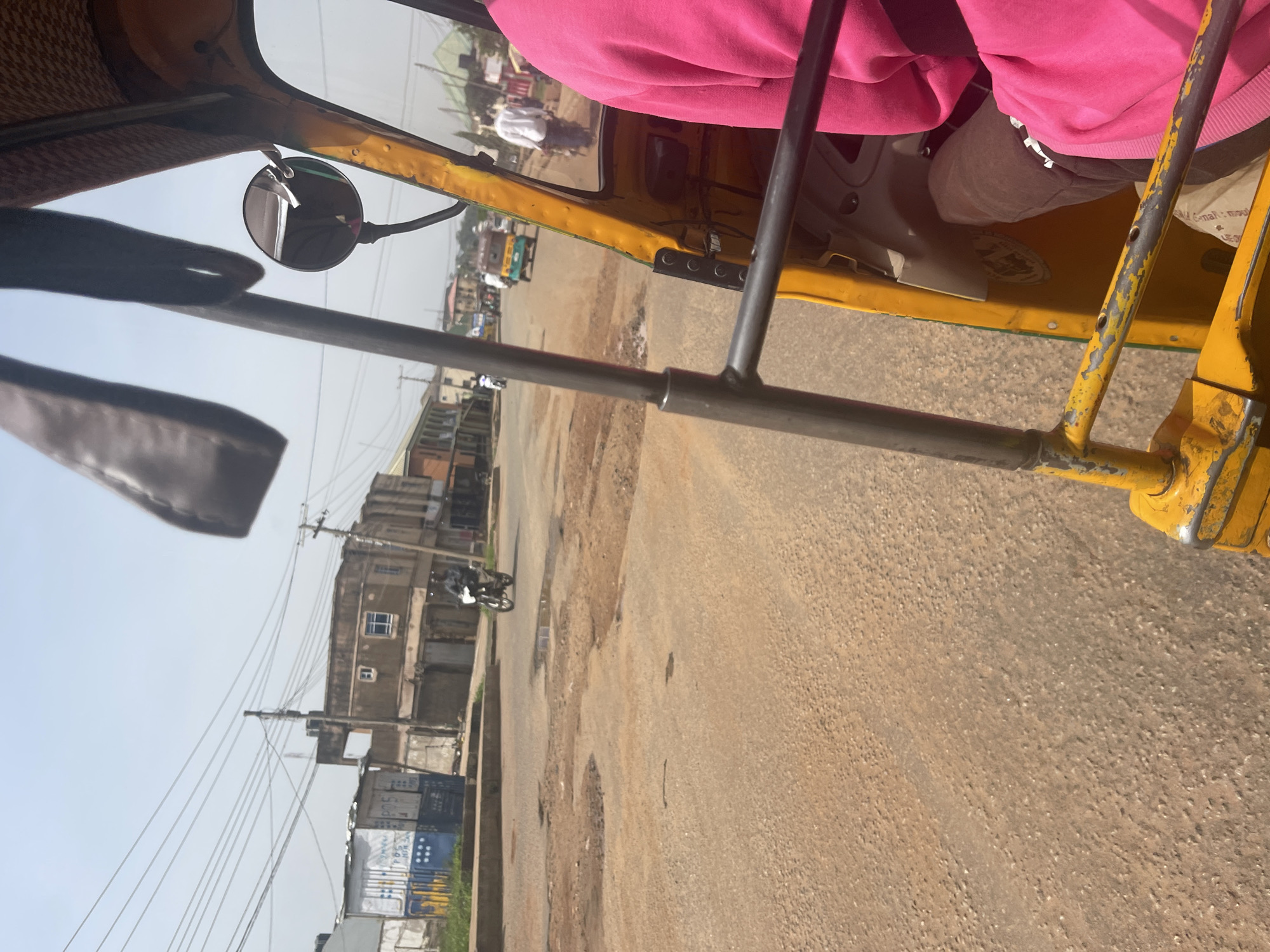 The image depicts a street scene in Rafin Par, Nigeria, viewed from the interior of a yellow auto-rickshaw. The rickshaw's metal frame and a passenger wearing a pink hoodie are visible in the foreground. The street is unpaved and appears to have several potholes. Buildings line the street, some appearing to be shops or residences. A motorcycle with a rider is seen on the road in the middle ground. Other vehicles, including what appears to be another auto-rickshaw, are visible further down the street. Overhead power lines crisscross the sky. The overall impression is of a typical urban street in Nigeria.