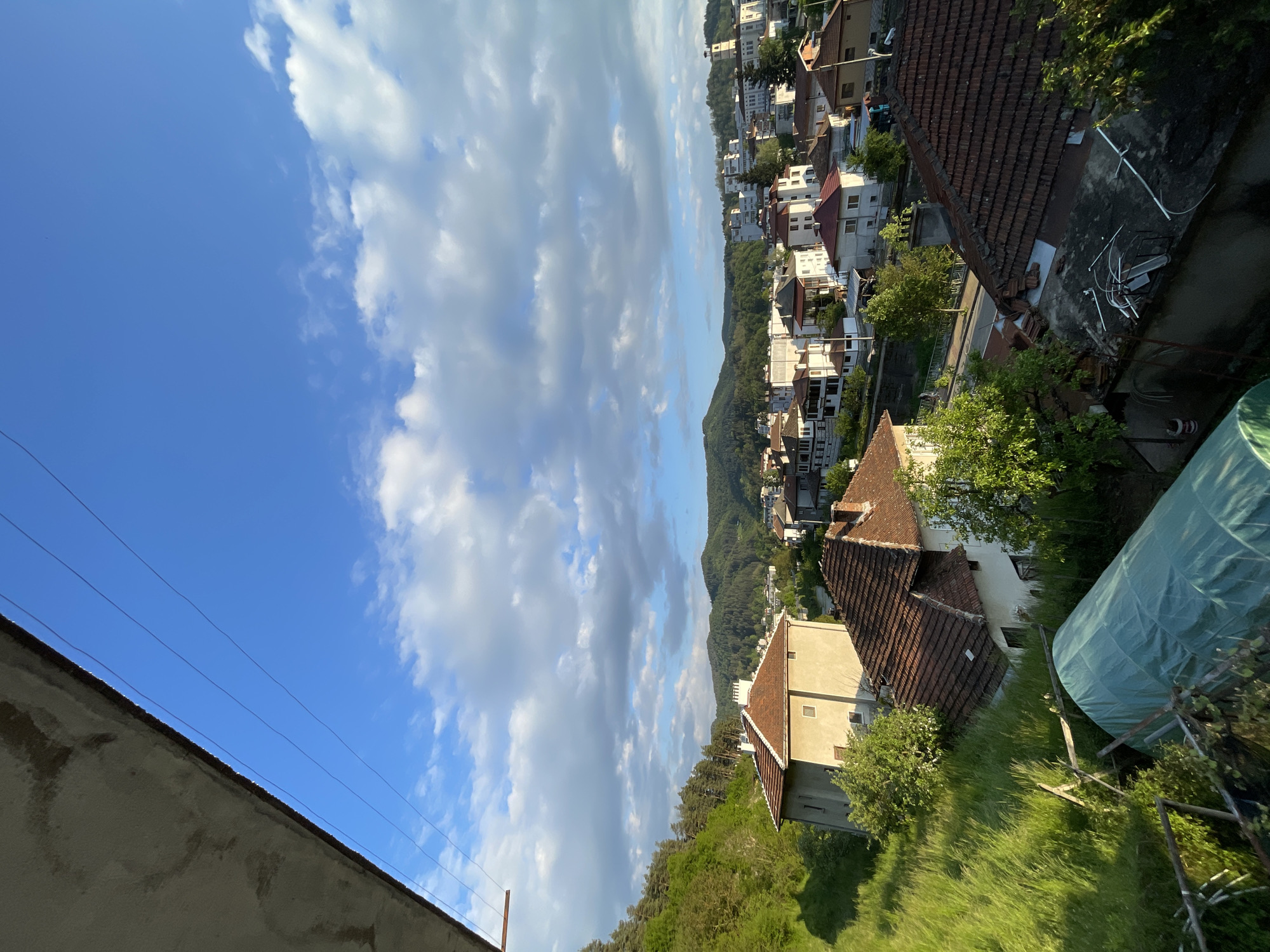 A slightly over-vertical shot captures a neighborhood in Zlatograd, Bulgaria under a partly cloudy, bright blue sky. The shot is taken from an elevated perspective. Rows of houses are visible, with their characteristic red-tiled roofs. Lush greenery and trees are interspersed amongst the houses and on the surrounding hills. A greenhouse can be seen on the right side of the image. The houses are mostly white. The environment is an urban residential area. There is no sign of humans. Overhead electrical wiring is seen in the lower left. The architecture and landscape seem to reflect a typical Southeastern European cityscape.