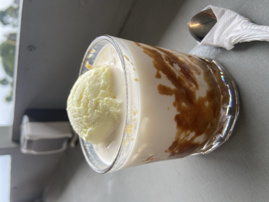 A dessert beverage is in a clear, ridged glass. The drink appears creamy white with streaks of dark brown, possibly caramel or coffee. A scoop of light yellow ice cream floats on top. A silver spoon wrapped in a white napkin sits beside the glass. The setting suggests a cafe or restaurant with a neutral-toned table surface and a partial view of the outside.