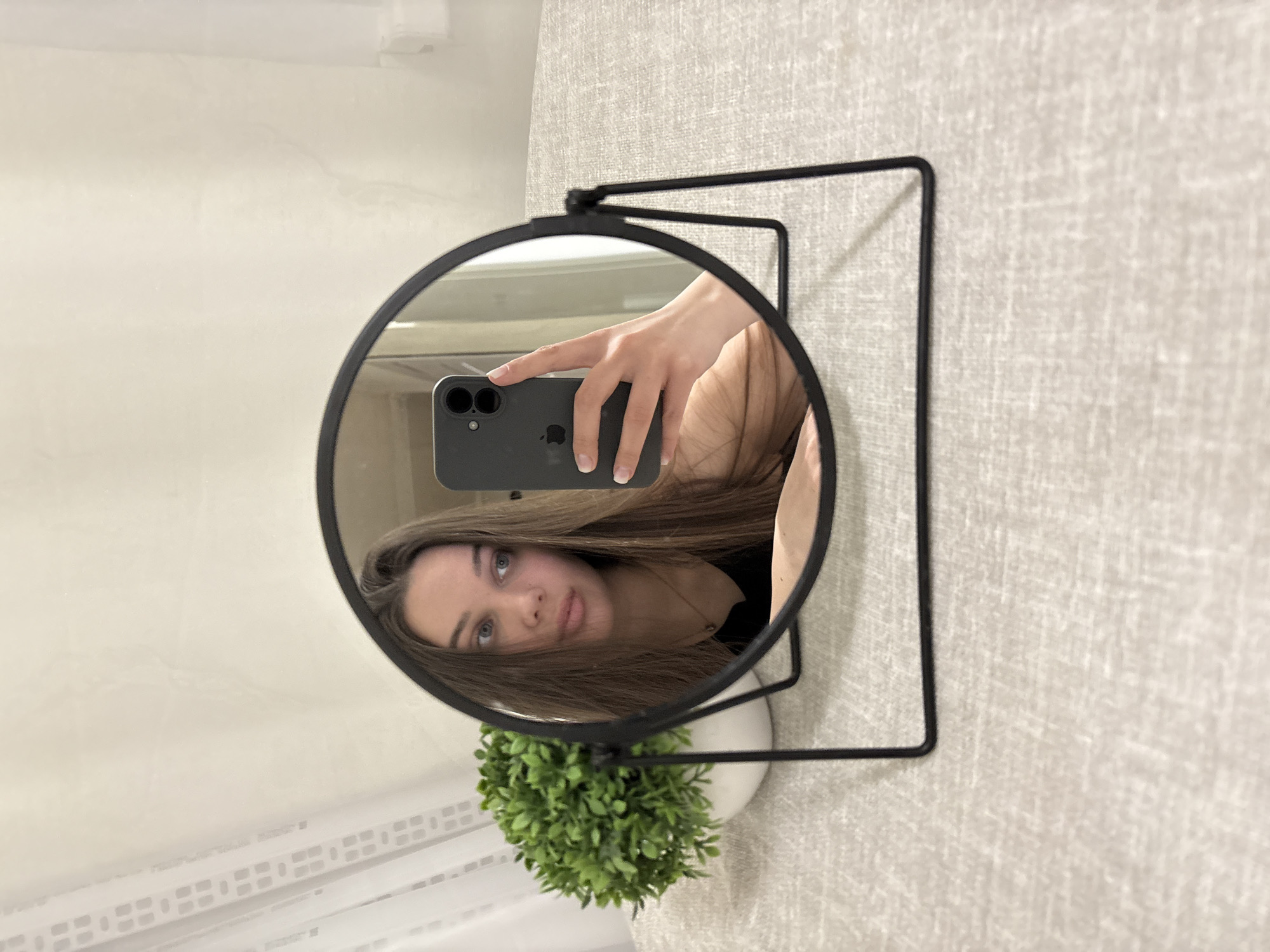An indoor scene in Chaikovsky, Russia, depicts a young female reflected in a round, black-framed tabletop mirror. The mirror is positioned centrally on a light grey, textured surface, likely a tablecloth. To the immediate left of the mirror, a potted artificial plant with vibrant green foliage in a white ceramic pot is visible. The reflection shows a female with long, straight brown hair, blue eyes, and a neutral expression, looking towards her left. She is wearing a dark-colored top and a gold-toned pendant necklace. Her right hand, with light-colored manicured nails, holds a dark grey smartphone (an iPhone model with dual rear cameras is identifiable by the Apple logo and camera configuration) directed towards the mirror, indicating a self-portrait (selfie) being taken. The background, visible behind the mirror, consists of a light-colored, textured wall. A sheer white curtain with a subtle grid pattern is partially visible on the left side of the background, and a white architectural element, possibly a window frame, is present on the right.