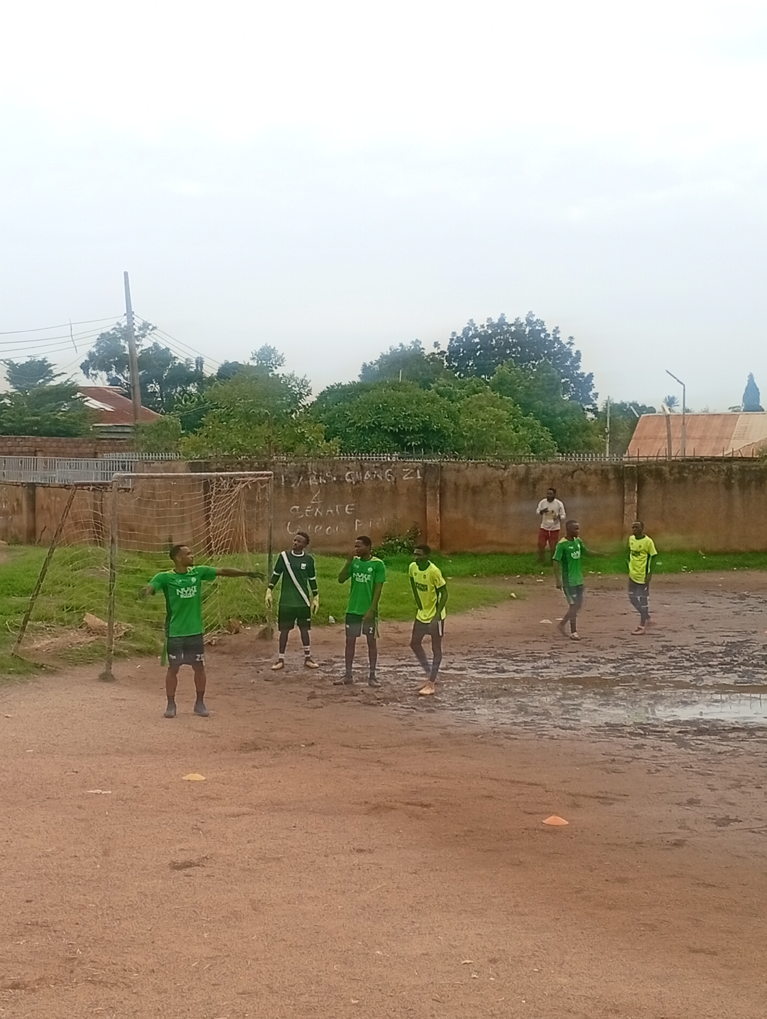 A group of young men in green athletic attire are on a muddy, dirt soccer field, possibly for practice. One man, wearing goalkeeper gloves and a green jersey with white accents, stands near a soccer goal. Another man in a green jersey and black shorts with the number 25 visible is positioned in front of the goal. To the right, two other men in green jerseys and black shorts are also on the field, one with the number 2. Further in the background, another man in a green jersey and dark pants walks, and a man in a white t-shirt and red shorts stands near a wall. The environment is an outdoor field with a rough surface, a soccer net, and a wall with writing on it that appears to be in English. Trees and buildings are visible in the background under a cloudy sky. The scene suggests an informal soccer training session in an urban setting.