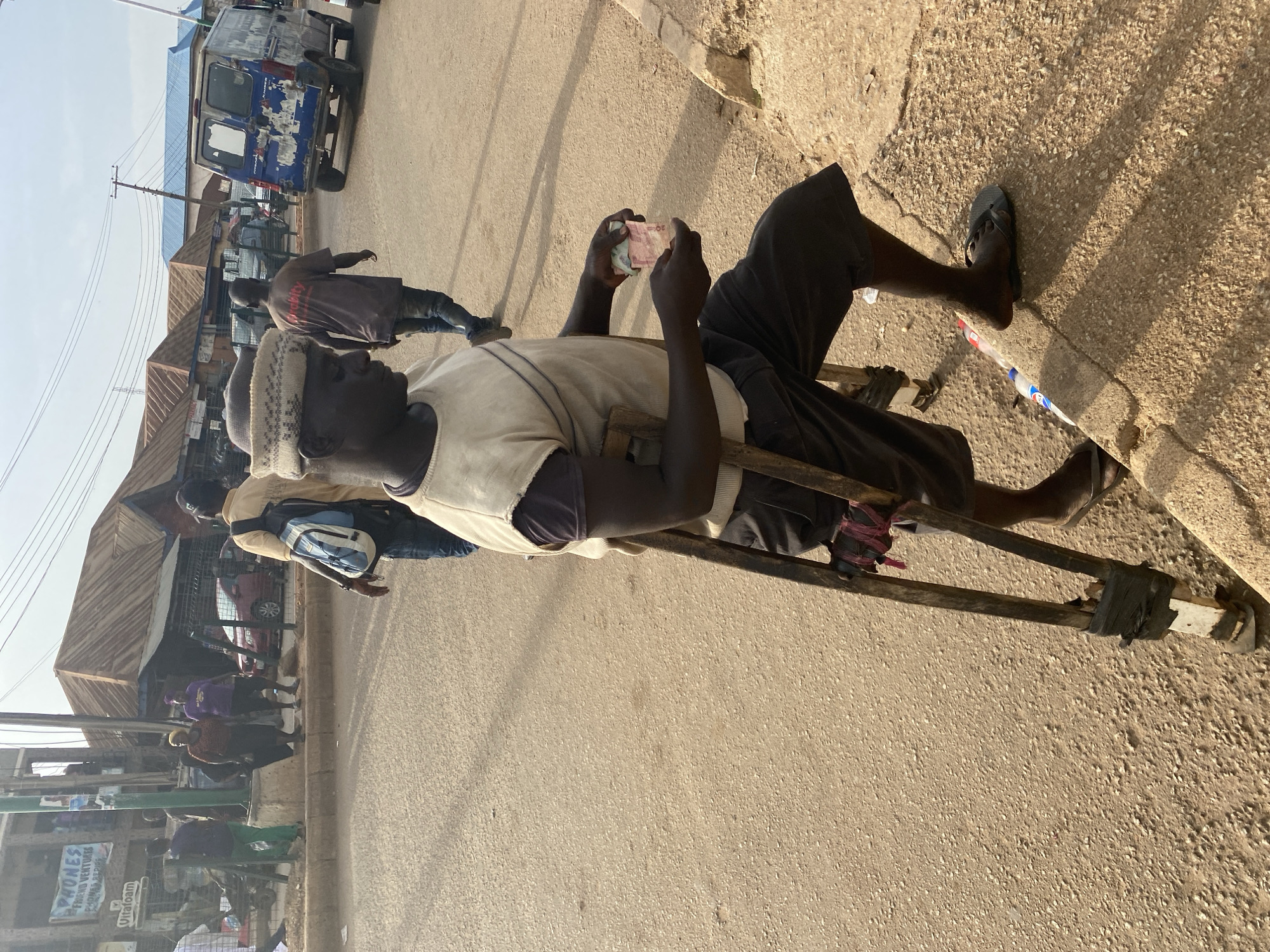 In Bukuru, Nigeria, a man wearing a knitted hat, a white vest, and black shorts sits on a wooden cart. He holds and examines currency in his hands. His feet are clad in dark flip-flops. The cart, seemingly makeshift, is made of wood and held together with what appears to be black tape. He is located on the side of a paved road. In the background, other people walk along the street amidst buildings and parked vehicles, including a blue van. Signage in the background indicates a commercial area.