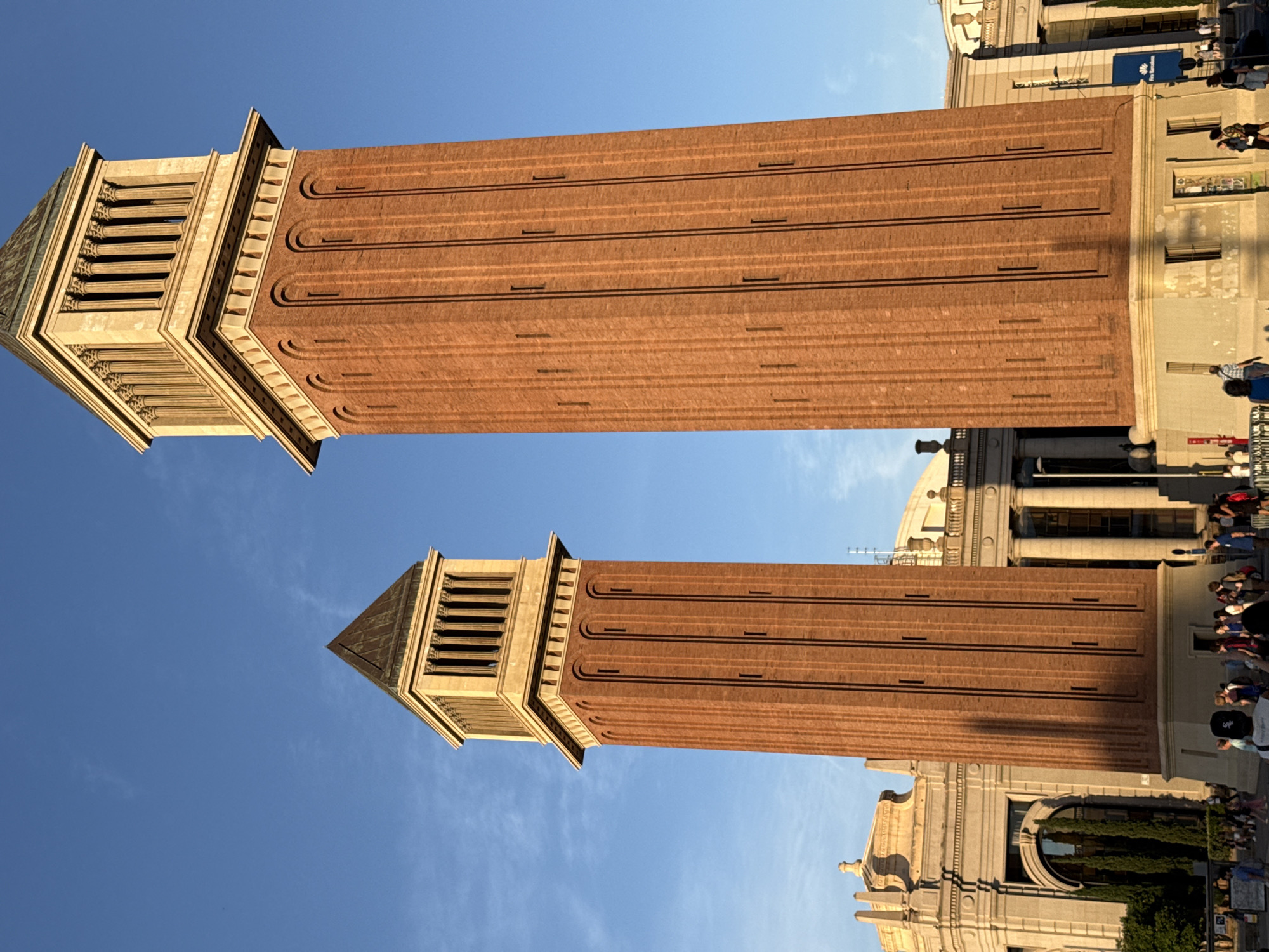 The image shows the Venetian Towers on Avinguda de la Reina Maria Cristina in Barcelona, Spain. The towers, constructed of brick with stone accents at the top, are positioned against a clear blue sky. The tower on the right is taller and closer to the viewer. In the background, classical-style buildings with columns and arched doorways are visible. A crowd of people is gathered in the foreground, walking and standing around the base of the towers. The lighting suggests it is late afternoon or early evening, with the sun casting long shadows. A blue banner with "Fira Barcelona" written on it is attached to one of the background buildings.