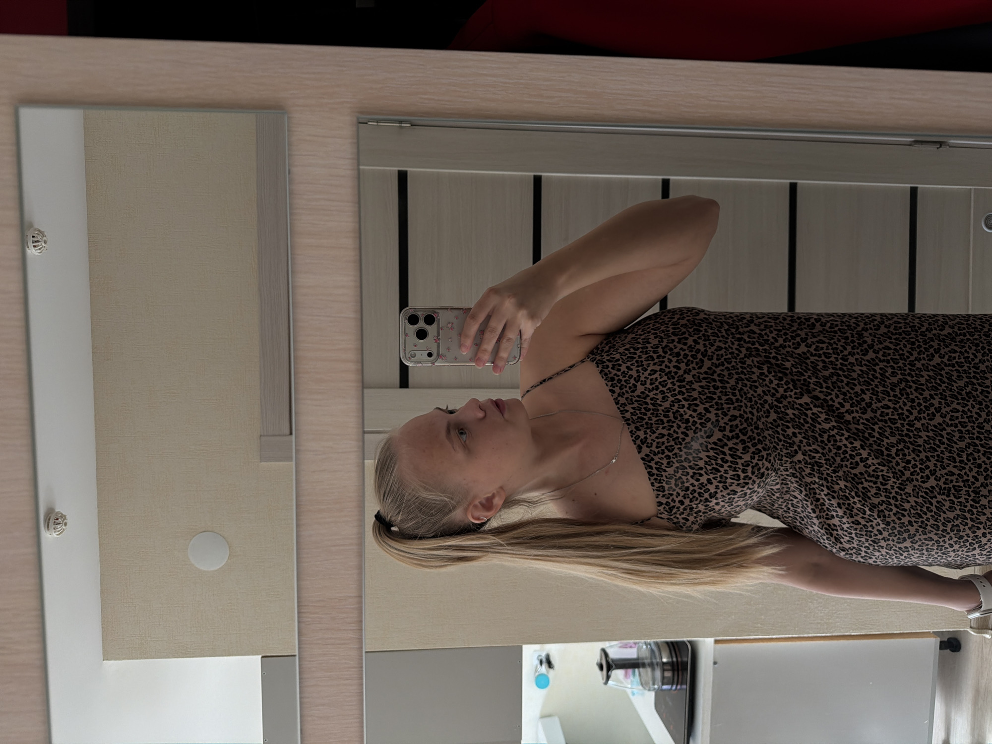 A blonde woman in a brown and black patterned dress takes a mirror selfie in what appears to be an indoor location. Her hair is pulled back in a ponytail. She's holding a phone with a clear floral case. The mirror reflects a door with vertical panels, a ceiling light, and what looks like a portion of a kitchen counter or appliance. The overall impression is that she is in a domestic setting. The location cannot be confirmed as Izhevsk, Russia from the visible details.