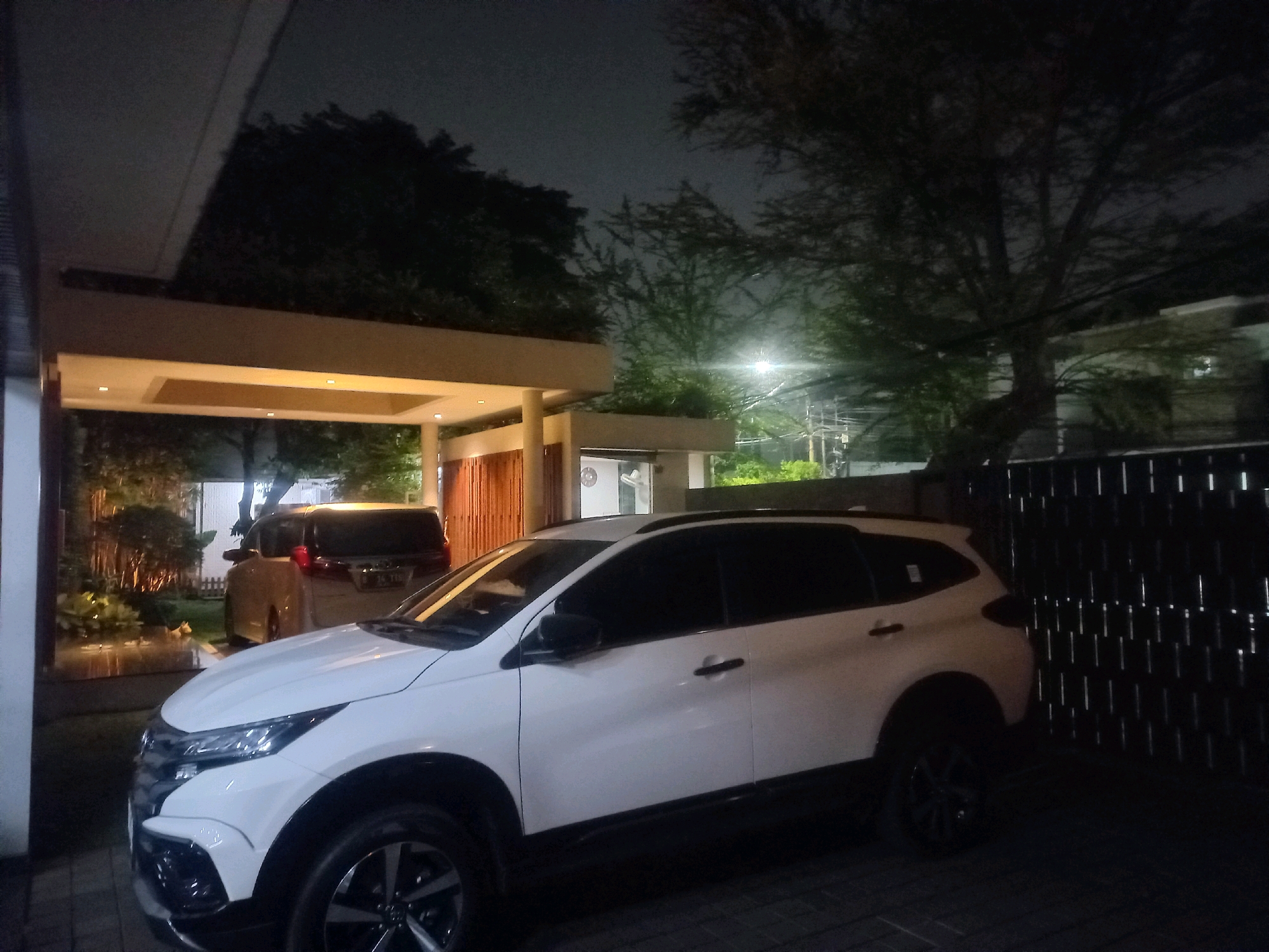 This is a photograph taken at night on a street in South Jakarta, Indonesia. The scene features two cars parked under a covered carport. The foreground car is a white SUV, partially obscuring a darker colored SUV parked behind it. The carport has a beige-colored overhang with embedded lighting. Beyond the carport, trees and foliage are visible against the dark night sky. A fence made of vertical metal bars is situated to the right of the scene. Electrical poles and wires can be seen in the background, indicating an urban setting. The lighting in the image is dim, primarily coming from the carport lights and ambient streetlights.