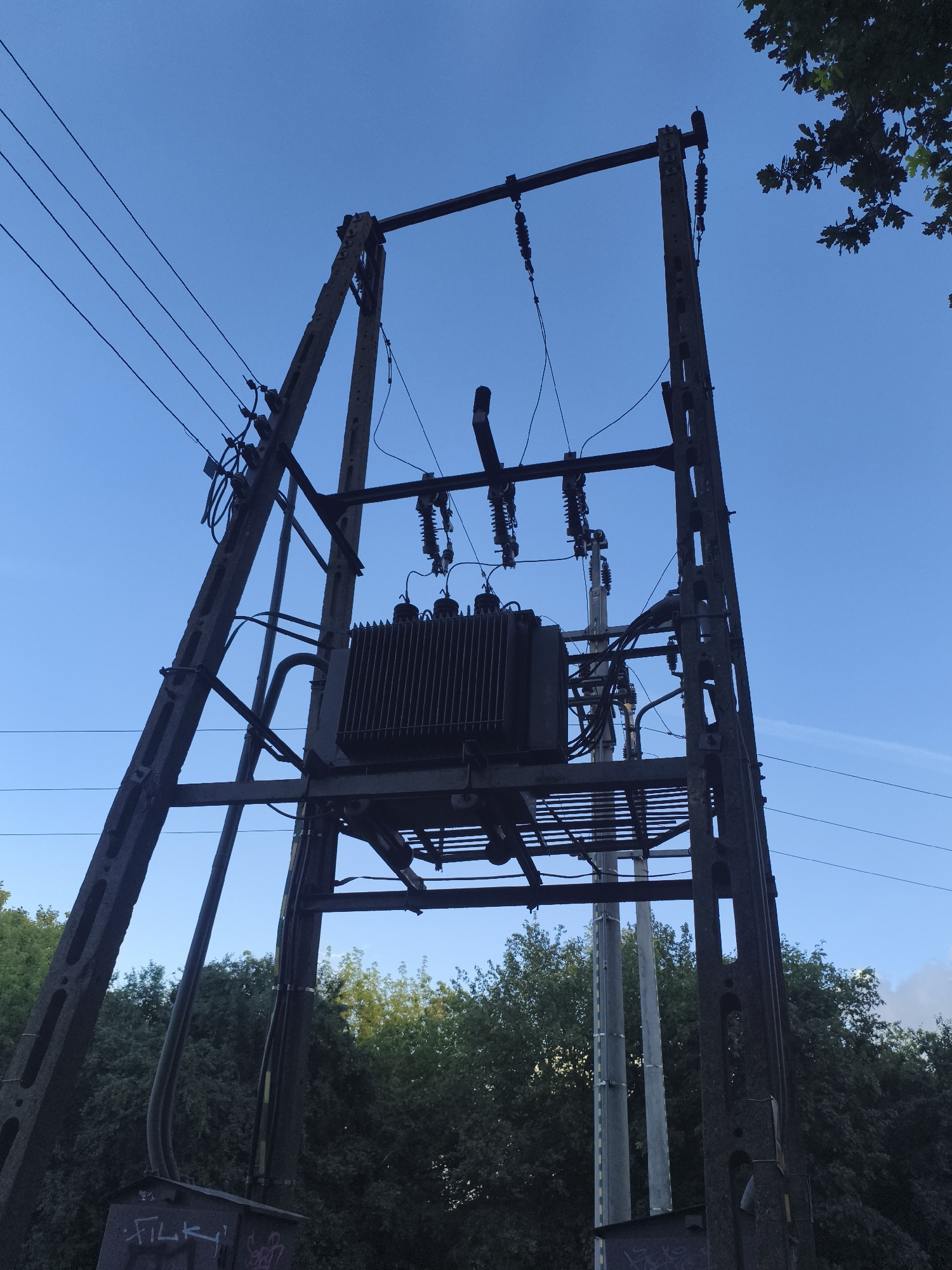 The image shows a large electrical transformer mounted on a metal pylon against a clear blue sky. The transformer is dark gray and ribbed, with several electrical connections visible. The pylon is constructed from dark metal beams and is supporting multiple overhead power lines. Trees with lush green foliage are visible in the background, and a small portion of another pole or structure can be seen behind the main pylon. The image appears to be taken from a low angle, looking upwards towards the transformer and sky. The scene is industrial and functional, typical of an outdoor electrical substation. There is no indication of the specific location within Nowy Dwór Mazowiecki, Poland, beyond the general context of an urban or semi-urban environment with electrical infrastructure.