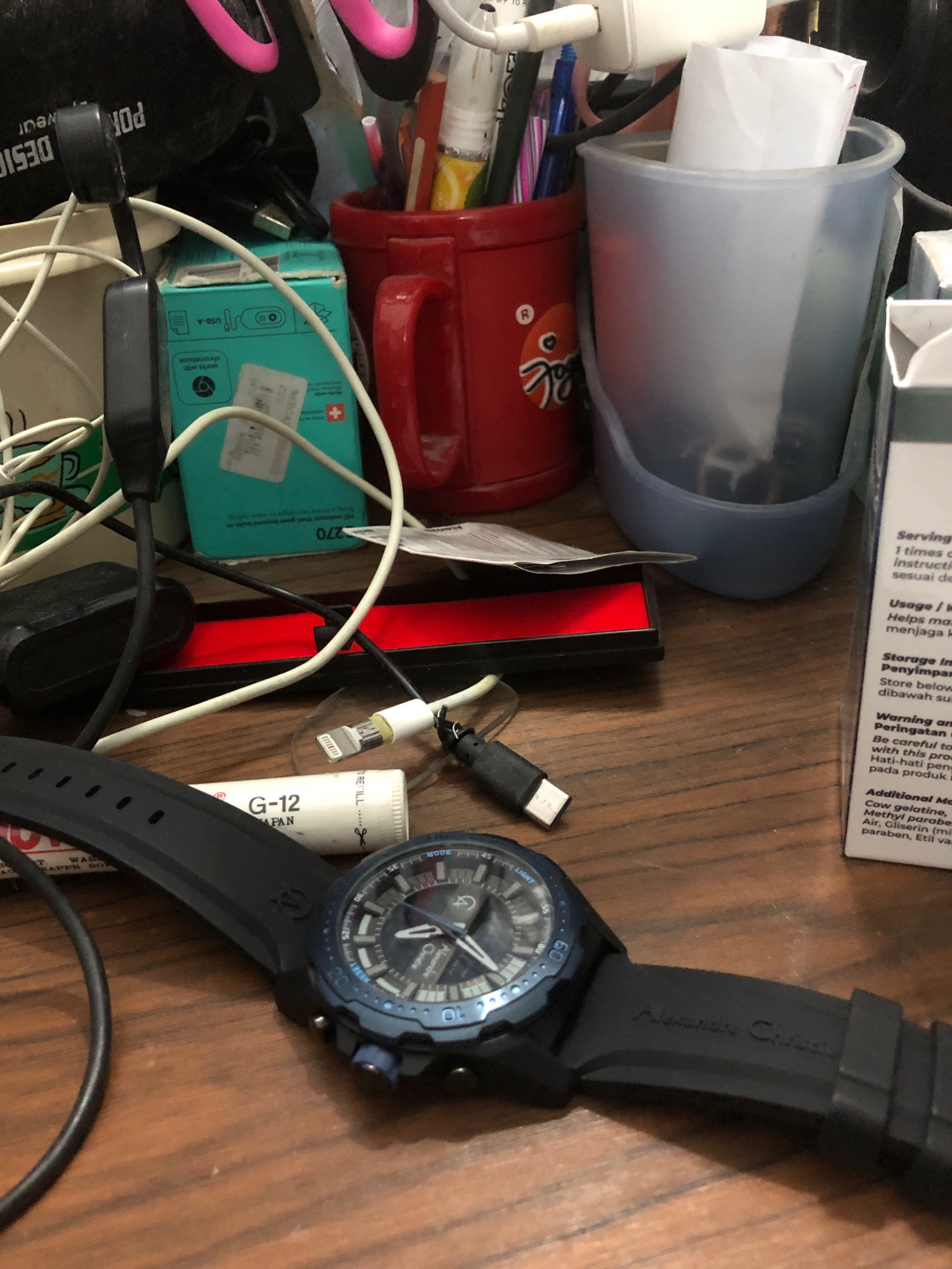 The image shows a cluttered wooden table with various electronic accessories, office supplies, and a watch. On the table, there is a black watch with a blue dial and the brand name "Alexandre Christie" inscribed on its strap. Nearby, a white tube labeled "G-12 JAPAN" is visible, along with a USB charging cable with a white connector. Other objects include a red mug, a blue plastic cup filled with items, a turquoise box, and several other charging cables and adapters. A white box with text in English and Malay is also present on the right side of the frame. The overall scene suggests a personal workspace or a desk area with everyday items. There are no indications or clues within the image to suggest its location in the city of Lhokseumawe, Indonesia.