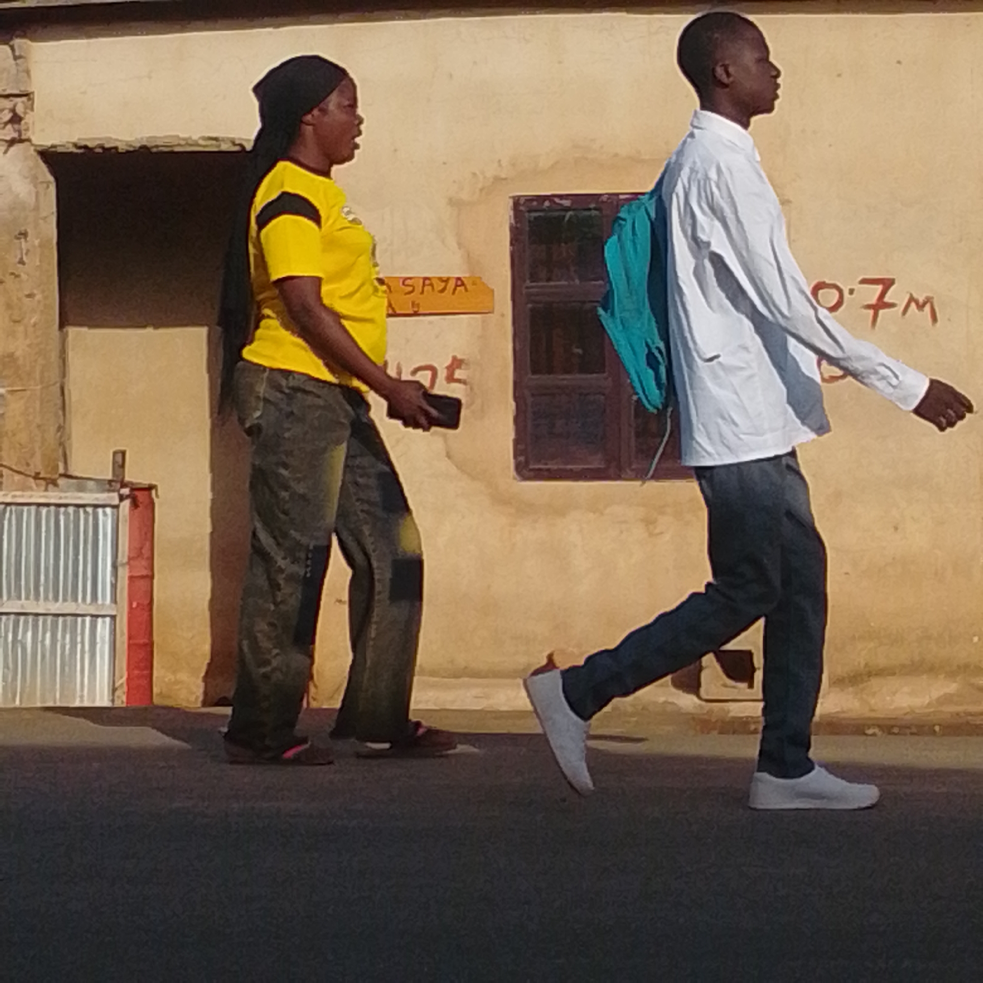 In Tempure, Nigeria, two individuals are walking on a paved surface in front of a textured, light-colored building. On the left, a woman wearing a black headscarf and a bright yellow t-shirt with black accents is walking to the right. She is wearing dark, loose-fitting pants with patches of lighter fabric. She is holding a dark rectangular object, possibly a phone, in her right hand. To her right, a young man is walking to the right, wearing a white long-sleeved shirt and dark pants. He has a teal backpack slung over his left shoulder. The building behind them has a window with dark brown trim and graffiti or writing in red on its surface, including "0.7M". To the far left, a corrugated metal structure with a red barrel next to it is visible. The scene is illuminated by sunlight, casting shadows on the ground.