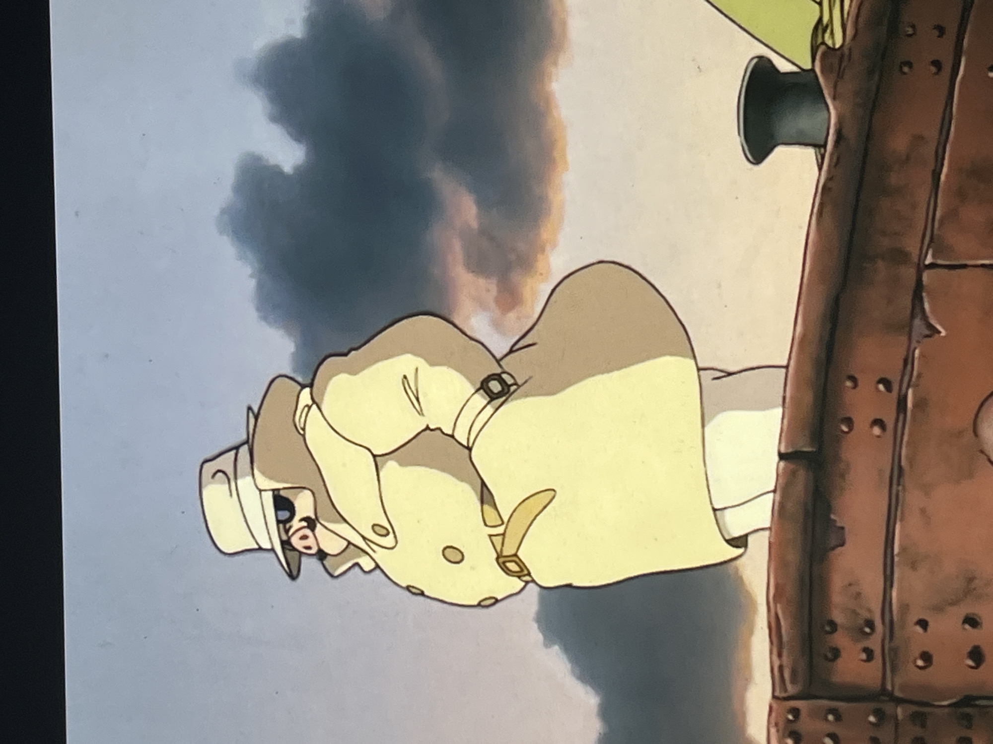 An animated still depicts an anthropomorphic pig character standing in profile on the upper structure of a vessel. The character wears a light-colored fedora, dark oval sunglasses, and a beige, double-breasted trench coat cinched with a matching belt. A watch is visible on its left wrist, and its hands are tucked into the coat pockets. The character looks towards its right. In the foreground, a riveted, reddish-brown metal structure, likely a boat's railing or hull, forms the base. To the far right of this structure, a dark, cylindrical bollard is present, alongside a partial view of a yellowish-green element, possibly a wing or sail. The background shows a sky with a light blue-grey upper portion, transitioning into a dramatic formation of dark, voluminous clouds, some exhibiting orange and pink hues, suggesting either dawn or dusk. Specific visual cues or landmarks identifying a location within the city of Budva, Montenegro, are not discernible in the image.