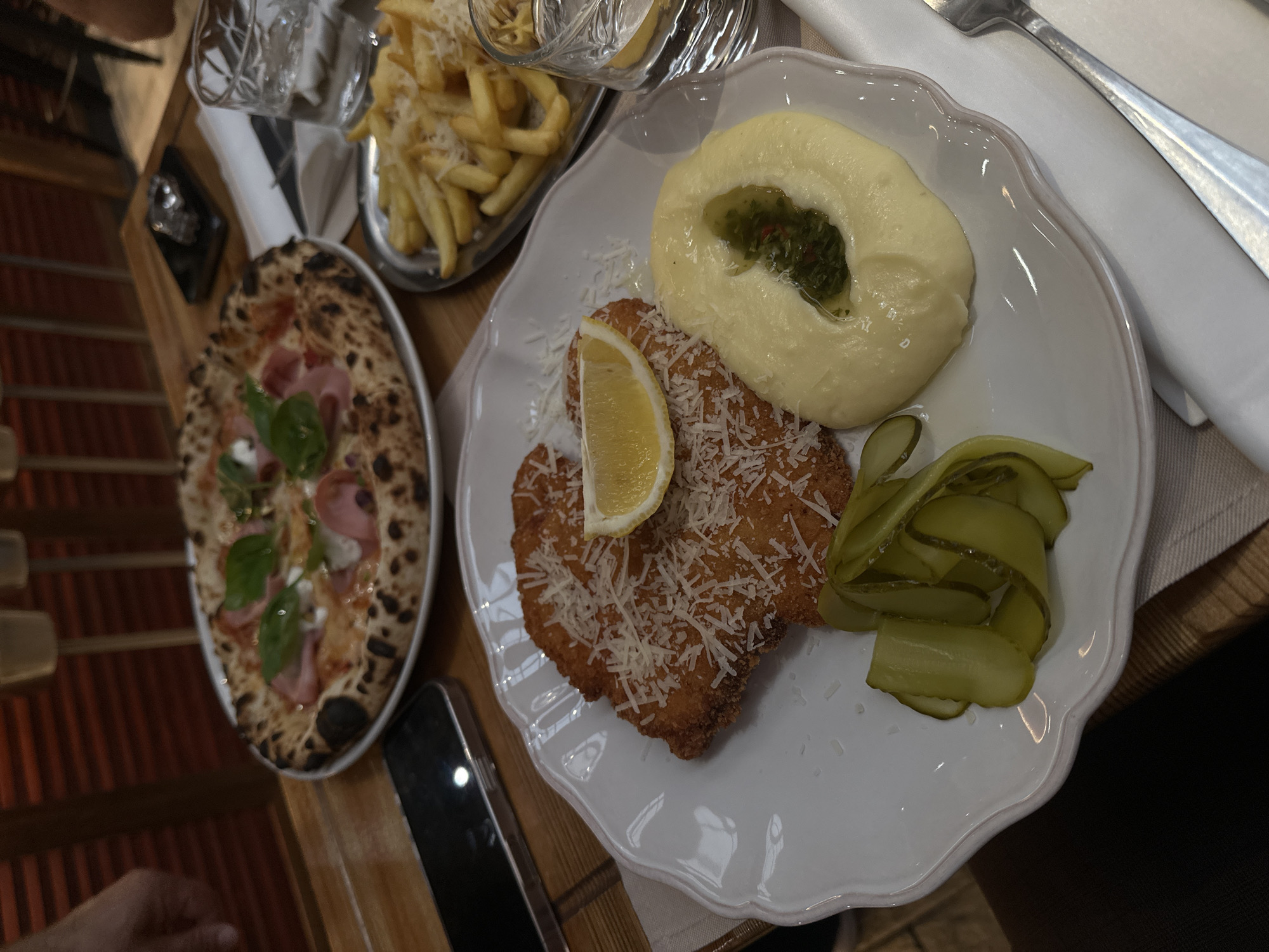 A top-down shot captures a table setting in what is likely a restaurant. Featured prominently are three plates: one with a pizza topped with basil, mozzarella, and ham; another holds french fries sprinkled with parmesan cheese; and a third plate contains what appears to be a breaded cutlet with a lemon wedge, a side of mashed potatoes with a green herb sauce in the center, and sliced pickles. A white linen napkin and cutlery are partially visible on the right. A dark-colored smartphone lies between the pizza and the plate with the cutlet, and the wooden tabletop is discernible beneath the plates. Based on the menu items and style of food preparation, this image might have been taken in a restaurant that serves a mix of European cuisines. There is no information present in this image to identify the city of Yerevan, Armenia, or any details beyond what's on the table.