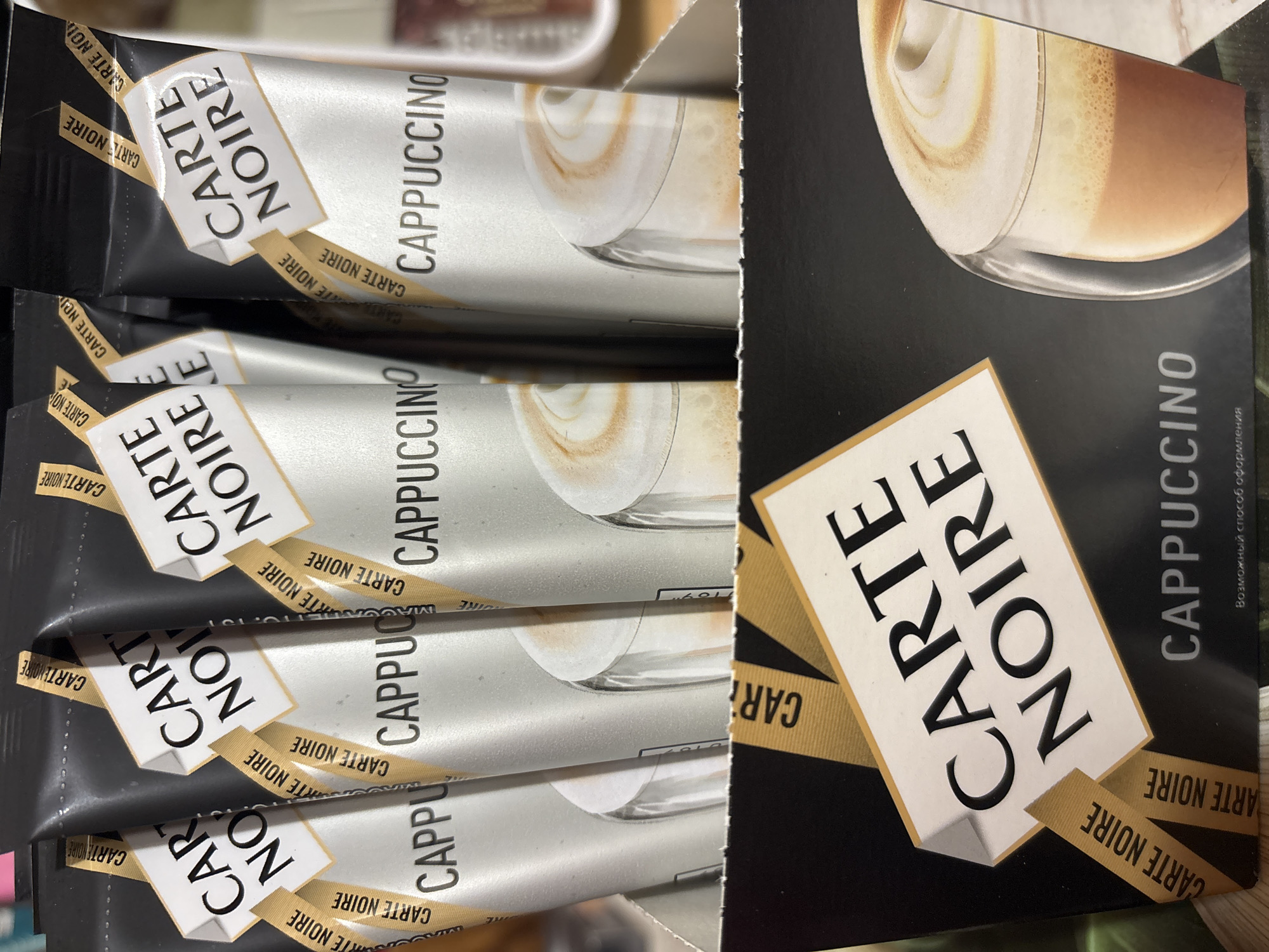 The image displays a retail product packaging for "Carte Noire Cappuccino". It features multiple pouches of instant coffee, each with the brand name "Carte Noire" and the product "Cappuccino" prominently displayed. The packaging is predominantly black with gold accents. The pouches show a graphic of a cappuccino. The bottom portion of the image reveals a box with the "Carte Noire Cappuccino" branding and a clear depiction of a prepared cappuccino in a glass. The product is likely for sale in a commercial setting, possibly a grocery store or supermarket. There is no information to suggest this image is related to Nadezhda, Russia.