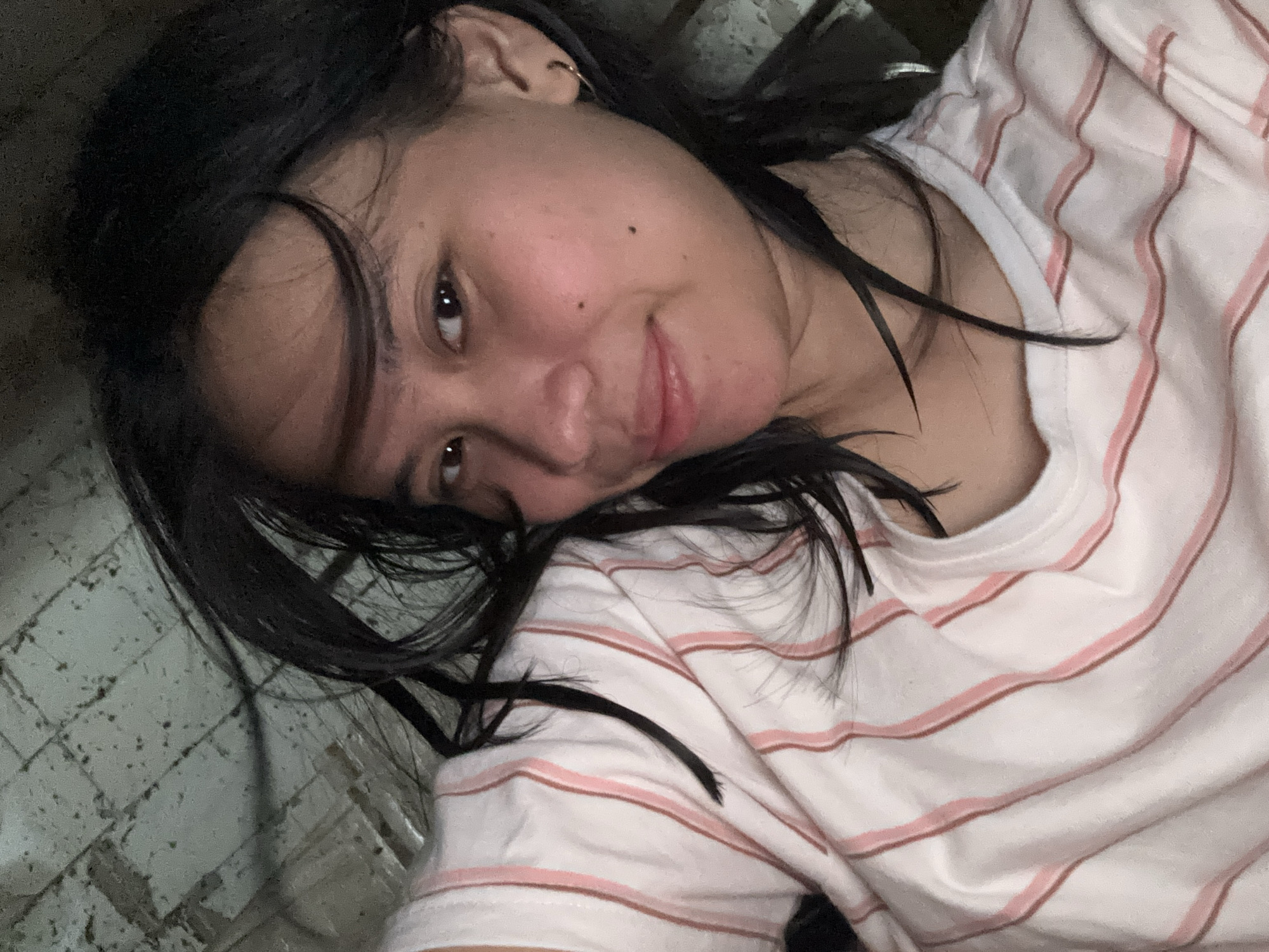 In this close-up, overhead shot, a young woman with dark hair and brown eyes smiles at the camera. She is wearing a white t-shirt with thin pink stripes. Her hair is tousled, with strands falling across her face and shoulders. The background appears to be a tiled floor, possibly in a residential setting in East Jakarta, Indonesia, though no specific landmarks are visible. The lighting is dim, suggesting an indoor environment. The overall impression is of a casual, personal moment captured candidly.