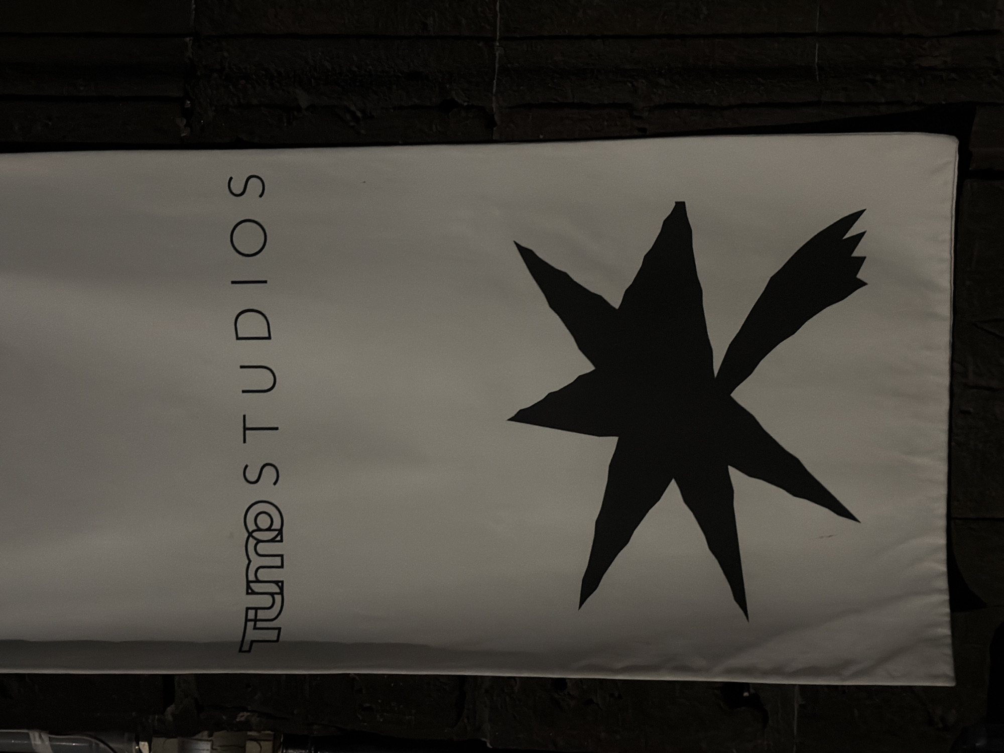 The image depicts a banner for "TUMO Studios" hanging against a dark, textured brick wall. The banner is predominantly white with black text reading "TUMO STUDIOS" vertically along the left side and a stylized, star-like graphic to the right. The environment appears to be dimly lit, emphasizing the contrast between the banner and the background. Based on the "TUMO Studios" branding, the location is likely in Yerevan, Armenia, as TUMO is an educational center primarily located there.