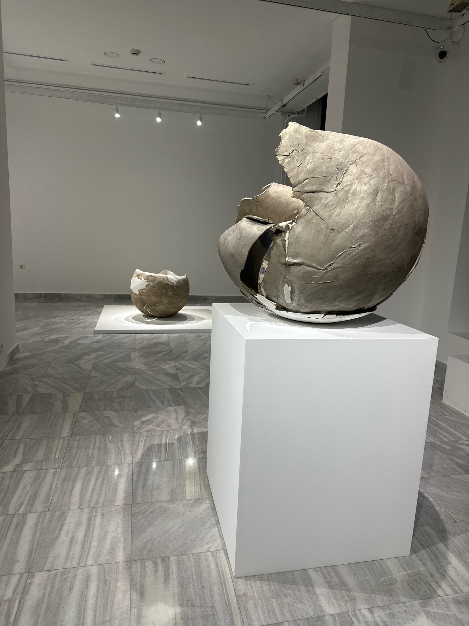 An indoor art exhibition space features two large, fragmented spherical sculptures. The foreground sculpture, positioned on a white cubic pedestal, is a significant size and appears to be made of a textured, light-colored material with cracked surfaces and visible internal structure. Behind it, a smaller, similar sculpture rests on a white rectangular platform, bathed in spotlighting. The room has white walls, a polished marble-tiled floor with a grey and white veined pattern, and modern track lighting visible on the ceiling. The environment suggests a contemporary art gallery or museum. This image was taken in Belgrade, Serbia.
