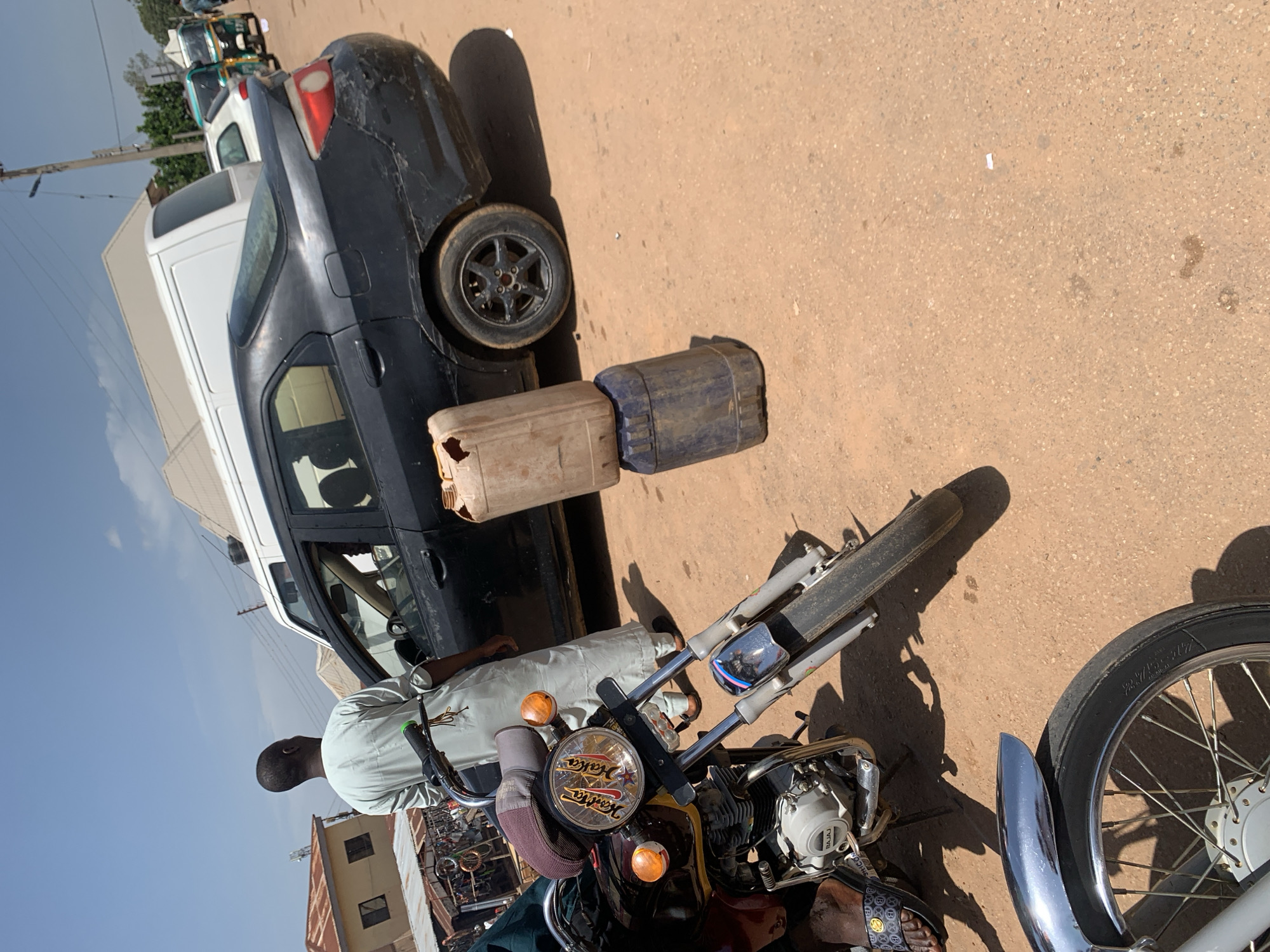The image captures an outdoor scene, likely a street or unpaved road, under bright daylight in what appears to be Gura Top, Nigeria. The strong shadows cast by vehicles and objects indicate a sunny, clear day, possibly mid-day or early afternoon.
In the foreground, a man is visible from the back, wearing a light-colored shirt, standing next to a motorcycle. He is wearing sandals with a patterned strap. The motorcycle, prominent in the lower half of the frame, has a round headlight with a decorative element that includes the words "Maska" and "Wakata." The engine casing also shows the brand name "BAJAJ."
Behind the motorcycle, a dark-colored car, possibly a sedan or hatchback, is parked. This car exhibits noticeable body damage and wear. Two large plastic jerrycans, one tan/orange and one blue, are placed on the ground beside the car, suggesting a potential activity related to fuel or liquid transfer.
Further in the background, other vehicles are visible, including a white van and a green and white tricycle (locally known as Keke Napep). A utility pole with wires runs vertically on the left side of the frame, and structures resembling buildings are present in the distance under a bright blue sky. The road surface is unpaved, appearing as dirt or gravel. The scene depicts a typical street environment with pedestrian and vehicular presence.