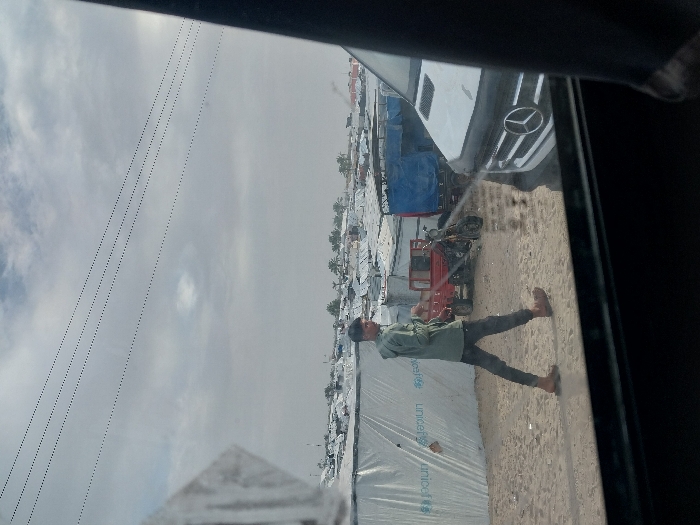 Through a vehicle window, a refugee camp stretches into the distance under a cloudy sky. Tents, some marked with "UNICEF," are prominent. In the foreground, a man in a light-green shirt walks on a dusty surface alongside the camp. A red motorized vehicle sits next to a blue tarp-covered structure. The front of a white Mercedes is visible through the window's reflection. Power lines run across the upper-left portion of the image.