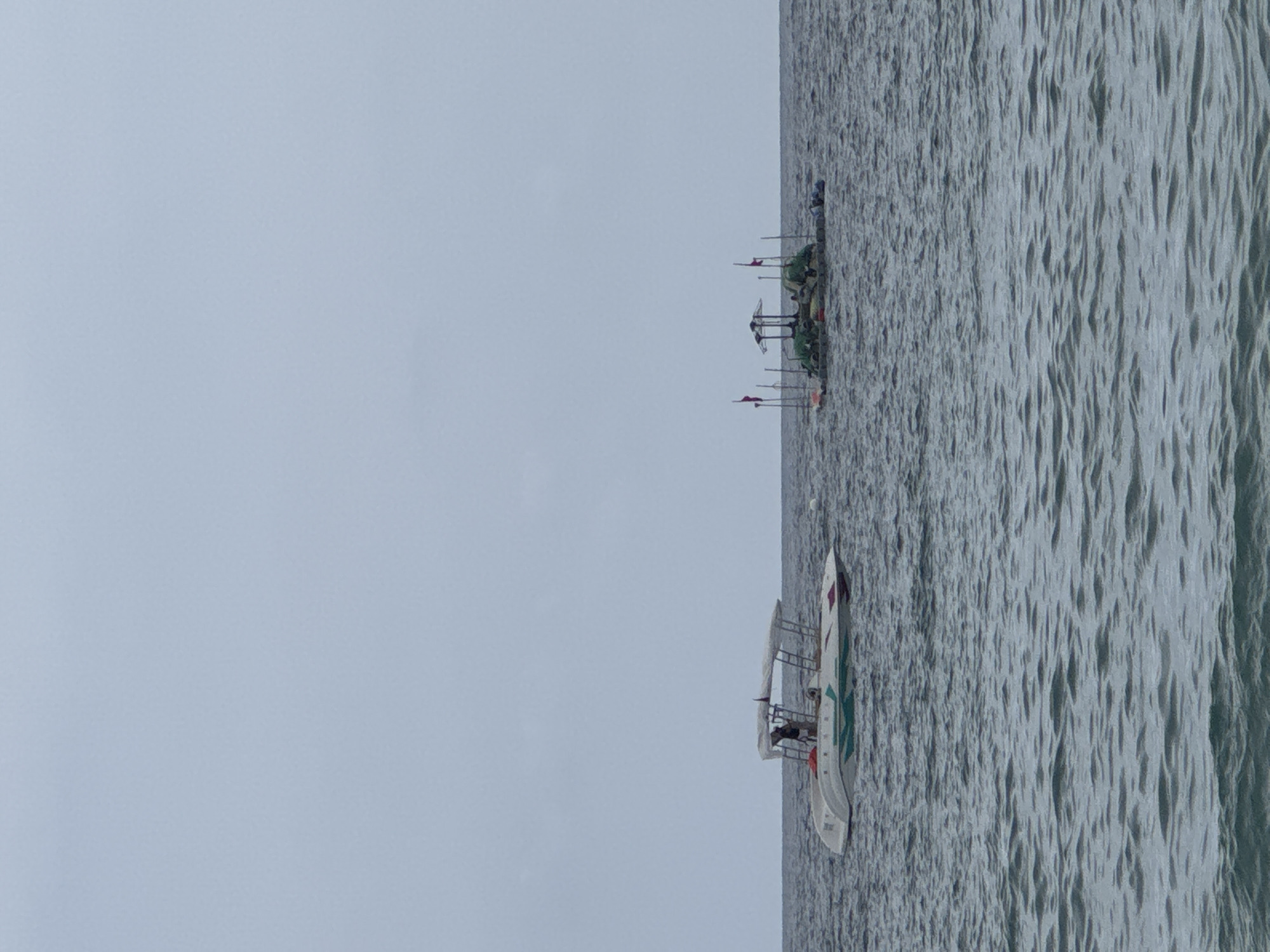 The scene captures a calm sea under a cloudy sky in Alanya, Turkey. Two vessels are visible: a trimaran and a fishing boat. The trimaran is a modern-looking, white boat with green and red accents. There are two people on the trimaran. The fishing boat is farther away, it is smaller, green and adorned with several flags. The water displays a light wave pattern, reflecting the dull light from the overcast sky. The overall atmosphere is serene and quiet, indicative of a day at sea.