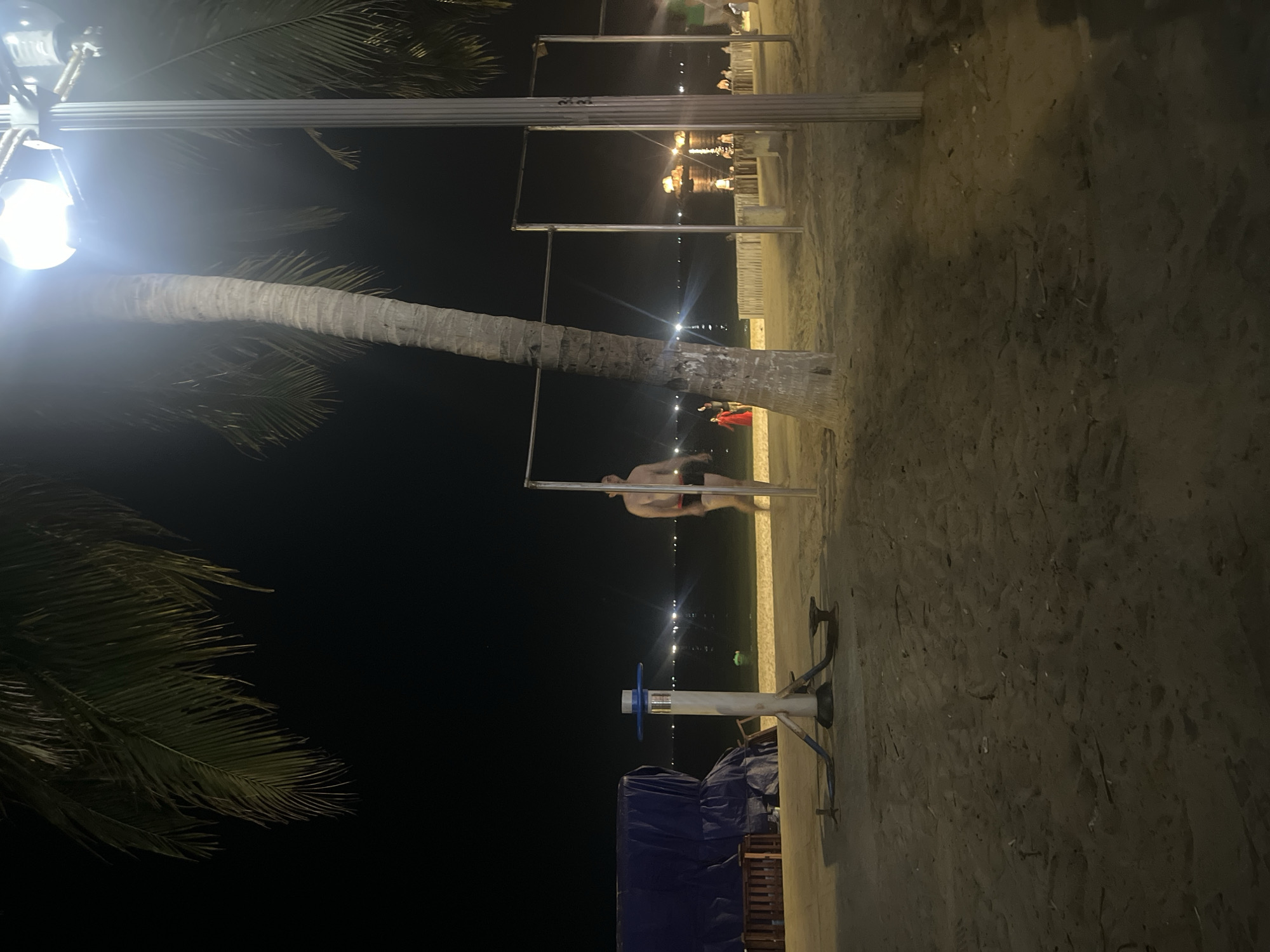 An outdoor nighttime scene on a sandy beach. A man wearing dark swim trunks stands to the right of a palm tree and appears to be walking away from the camera. In the background, a body of water is visible, dotted with distant lights. To the left, a blue tarp covers an object, and a piece of exercise equipment stands on the sand. A bright light pole with a lamp illuminates the scene.