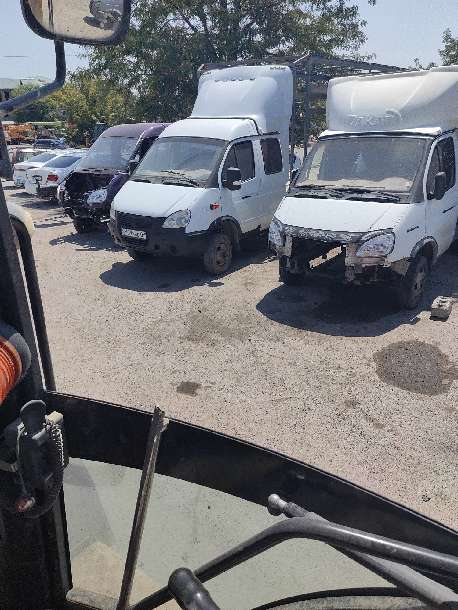 This is a horizontal, outdoor, eye-level, medium shot of a vehicle graveyard in Shymkent, Kazakhstan. The foreground is dominated by the interior of a vehicle, with part of a steering wheel visible on the left, along with some levers and a metal pole. The background shows several damaged or partially dismantled vehicles parked on a gravel lot. These include a purple van, a white van with a "DAKAR" logo on its top, and other white vans and cars. The vehicles appear to be in disrepair, with some missing parts such as bumpers and headlights. The environment is sunny, with trees visible in the distance and a clear sky overhead.