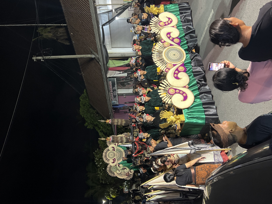 A nighttime street scene in Munggu, Indonesia, depicts a cultural or religious procession. The primary focus is a large, elaborately decorated float moving along an asphalt road. This float features a base draped in green and black fabric, adorned with intricate, light-colored, fan-like and swirling patterns, and two golden Garuda figures flanking small gongs. Behind it, a second, taller float displays similar decorative patterns and two prominent tall, dark, textured ornaments.
Approximately 30-40 individuals participate in the procession. Many wear uniform dark green long-sleeved shirts, black trousers, and traditional Balinese Udeng (head wraps). Some are positioned behind the main float, appearing to carry or attend to instruments. Others walk alongside, carrying white flags with black patterned edges on staffs.
In the foreground, several spectators observe the event from the side of the street. One individual, seen from behind in a pink shirt, holds a smartphone vertically, recording the procession. Another spectator in a black shirt and traditional headwear is positioned to their left. Part of a dark-colored vehicle is visible in the lower left corner.
The environment includes buildings lining the street, such as a structure with a brown tiled roof and a visible sign for "AFDAN GORDEN." A utility pole with power lines extends above the street. Artificial lighting illuminates the scene against a dark night sky. Palm trees and other foliage are visible in the background.