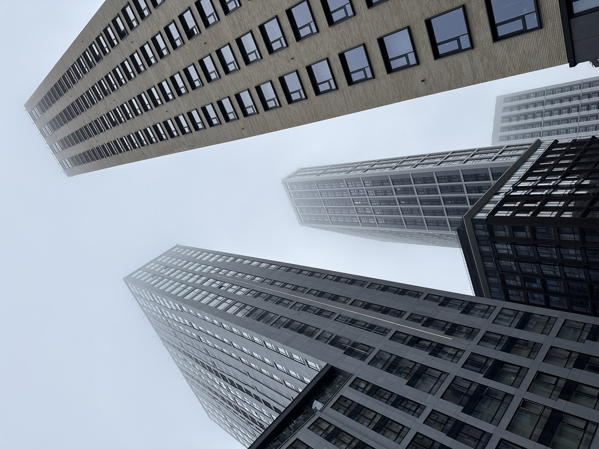 The image depicts a low-angle view of several tall buildings in Moscow, Russia. The buildings are of modern design with numerous windows and varying facade materials, including brick and metal. The sky is overcast and foggy, obscuring the upper portions of the buildings and reducing visibility. There are no visible people or indications of human activity. The focus is solely on the architecture and the atmospheric conditions.