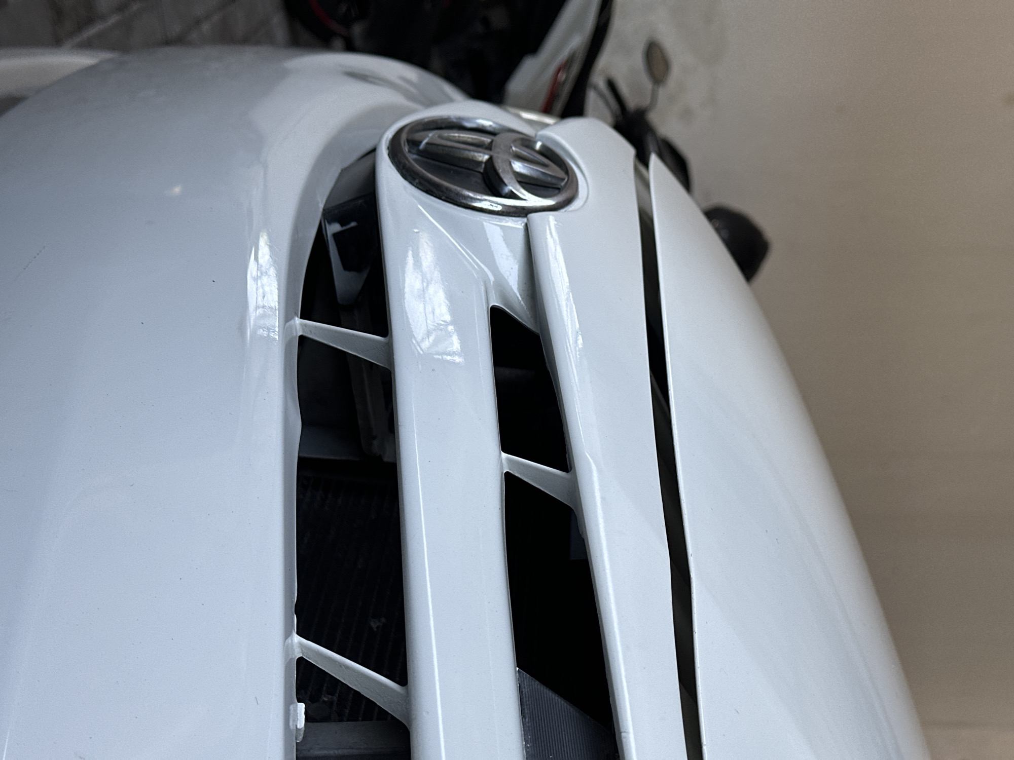 This is a close-up, low-angle, outdoor photograph of the front of a white car, likely a Toyota given the visible emblem. The car is positioned in what appears to be a garage or covered area, with a concrete floor and a textured, light-colored wall in the background. To the left of the car, partially visible, is a black motorcycle with red accents. The car's grille features horizontal white slats and a central chrome emblem. The lighting suggests it is daytime, possibly with ambient light filtering in. The image is focused on the front bumper and grille area of the vehicle. There are no people present in the scene. The location is specified as Bulakrejo, Indonesia.