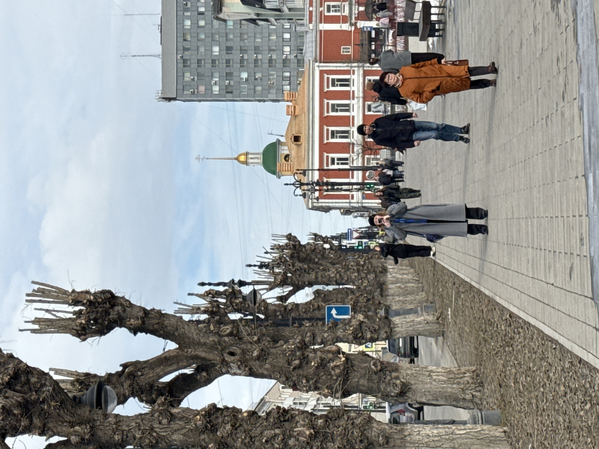 The image depicts a street scene in the city of Perm, Russia. A row of bare-branched trees lines the sidewalk. Pedestrians are walking along the sidewalk, including individuals wearing a brown coat, a black coat, and a gray coat. Buildings are visible in the background, including a modern high-rise and a traditional red brick building with white trim and a green-domed church. Streetlights are visible along the sidewalk. The sky is overcast with patches of blue.