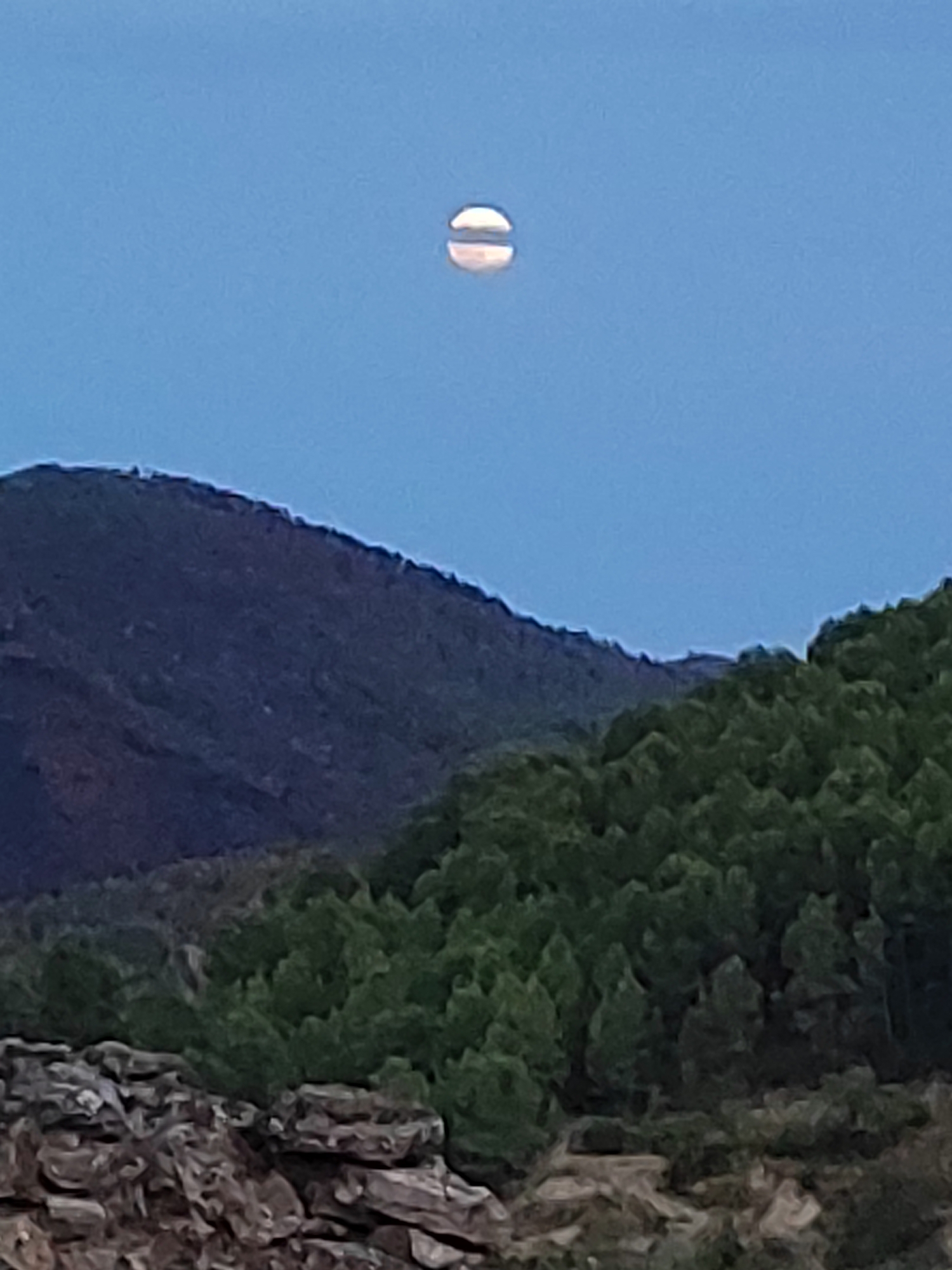 A full moon is visible in a clear blue sky above a mountainous landscape. The moon appears slightly hazy with darker shadows on its surface. The mountains in the foreground are covered with dense green trees. In the lower part of the image, there is a rocky embankment. The image appears to have been taken at dusk or dawn. The location is identified as Suera, Spain.