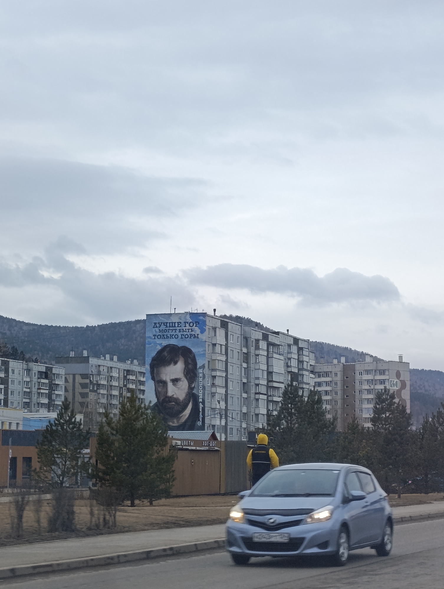 The image shows a cityscape, likely in Krasnoyarsk, Russia, given the Cyrillic script on the buildings. The sky is overcast with gray clouds. In the background, there are apartment buildings and a tree-covered hill. A large mural of a bearded man, presumably Vladimir Vysotsky, is painted on the side of one of the buildings, accompanied by Cyrillic text reading "Лучше гор могут быть только горы" which translates to "Only mountains can be better than mountains" . In the foreground, a blue Toyota Vitz (likely a Japanese import) is in motion on a road. To the side of the road, a pedestrian wearing a yellow hooded jacket is present. Also visible are several evergreen trees and a row of brown buildings containing a shop (flowers and hot dogs). The Cyrillic text "ЦВЕТЫ" and "ХОТ-ДОГ" can be read on a sign attached to the building.