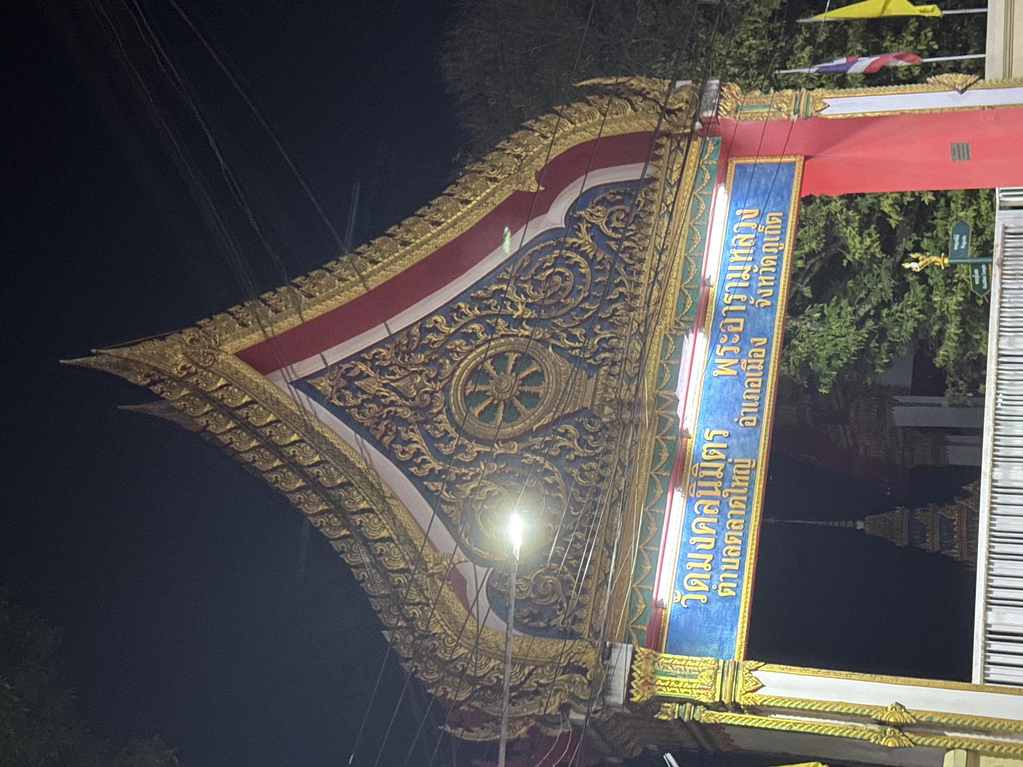The image shows a brightly lit, ornate temple entrance at night. The architecture is distinctly Thai, with a tiered, pointed roof decorated with intricate gold carvings against a dark blue background. The entrance features a large blue sign with Thai lettering, which appears to be the name of the temple and its location. The Thai text on the sign reads "วัดมงคลนิมิตร ตำบลตลาดใหญ่ อำเภอเมือง จังหวัดภูเก็ต," which translates to "Wat Mongkolnimit, Talat Yai Subdistrict, Mueang District, Phuket Province." This indicates the location is within the Mueang District of Phuket Province, specifically in the Talat Yai Subdistrict, which is part of the Phuket City Municipality. Two flags, one Thai flag and one yellow flag, are visible on the right side of the entrance. In the background, trees and the dark night sky are visible, along with the spire of another temple structure. The overall scene depicts a religious landmark illuminated at night, showcasing its detailed craftsmanship.