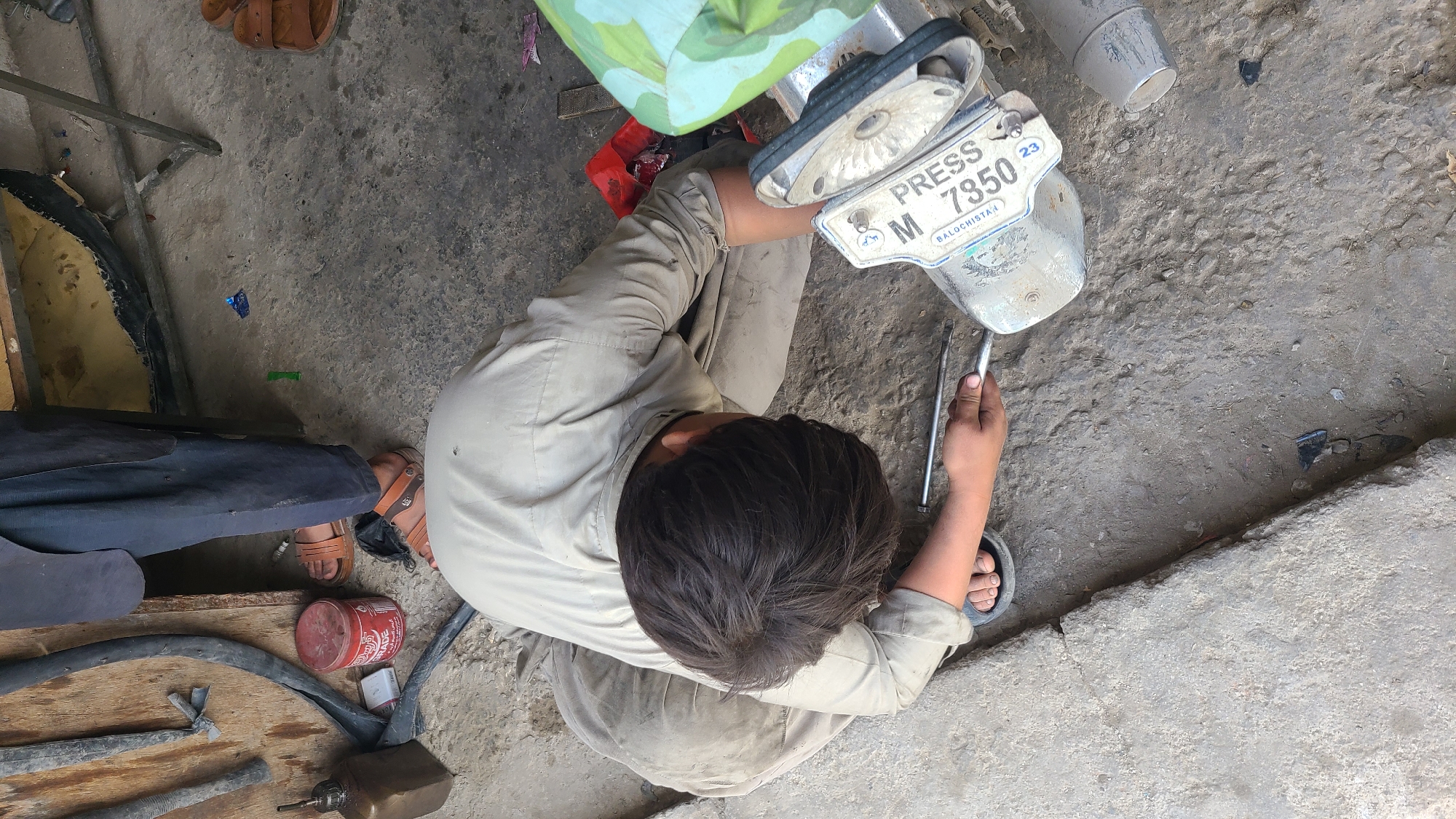 An overhead shot captures a person, likely a male mechanic, crouched on an uneven concrete or dirt surface, deeply engrossed in repairing a motorcycle part. He is wearing a light-colored (possibly beige or khaki) shirt and trousers, with dark slippers on his feet. His back is to the camera, showing the top of his head as he bends over the task.
In his right hand, the man holds a metal tool, possibly a wrench or screwdriver, using it on a silver-colored metallic component. This component appears to be a motorcycle's front headlight or mirror assembly, with a white license plate attached to it. Another similar tool rests on the ground beside his hand.
The visible text on the license plate reads "PRESS" at the top, "M 7850" in the middle, and "BALOCHISTAN" at the bottom, accompanied by a small "23" in the upper right corner. This indicates the vehicle belongs to a press organization and is registered in the Balochistan province of Pakistan, which aligns with the given location context of Quetta City Tehsil.
The setting suggests an outdoor or informal workshop. The ground is rough and dusty, with various items scattered around, including a red metal can, pieces of wood, and what look like discarded rubber or tire parts. The scene is brightly lit by natural daylight, indicating clear weather. The partial view of another person's legs and brown sandals in the lower left corner suggests others are nearby. The overall impression is one of practical, manual repair work taking place in an accessible, open environment.