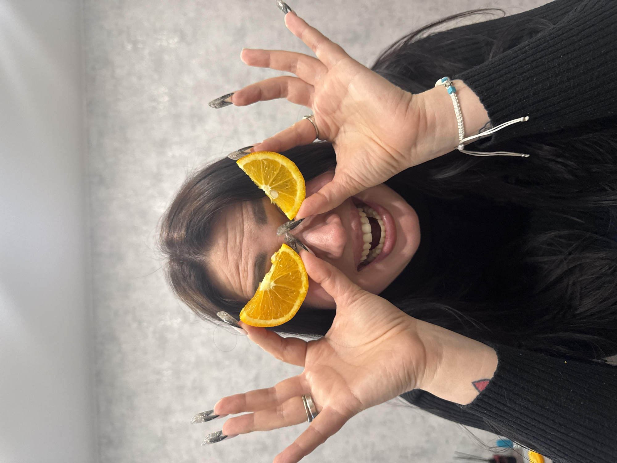 A playful, close-up, vertical shot shows a person, likely female, with long dark hair, holding two orange slices over her eyes. Her mouth is wide open in an exaggerated, happy expression, revealing teeth and tongue, and causing visible wrinkles on her forehead. She is wearing a black long-sleeved top. Her hands, adorned with long, pointed, silver glittery fingernails and silver rings, frame her face. On her left wrist, she wears a white woven bracelet with small blue beads. A small red triangle tattoo is visible on her left inner wrist, just peeking out from under her sleeve. The background is a plain, light gray textured wall, suggesting an indoor setting. There is no visible text.