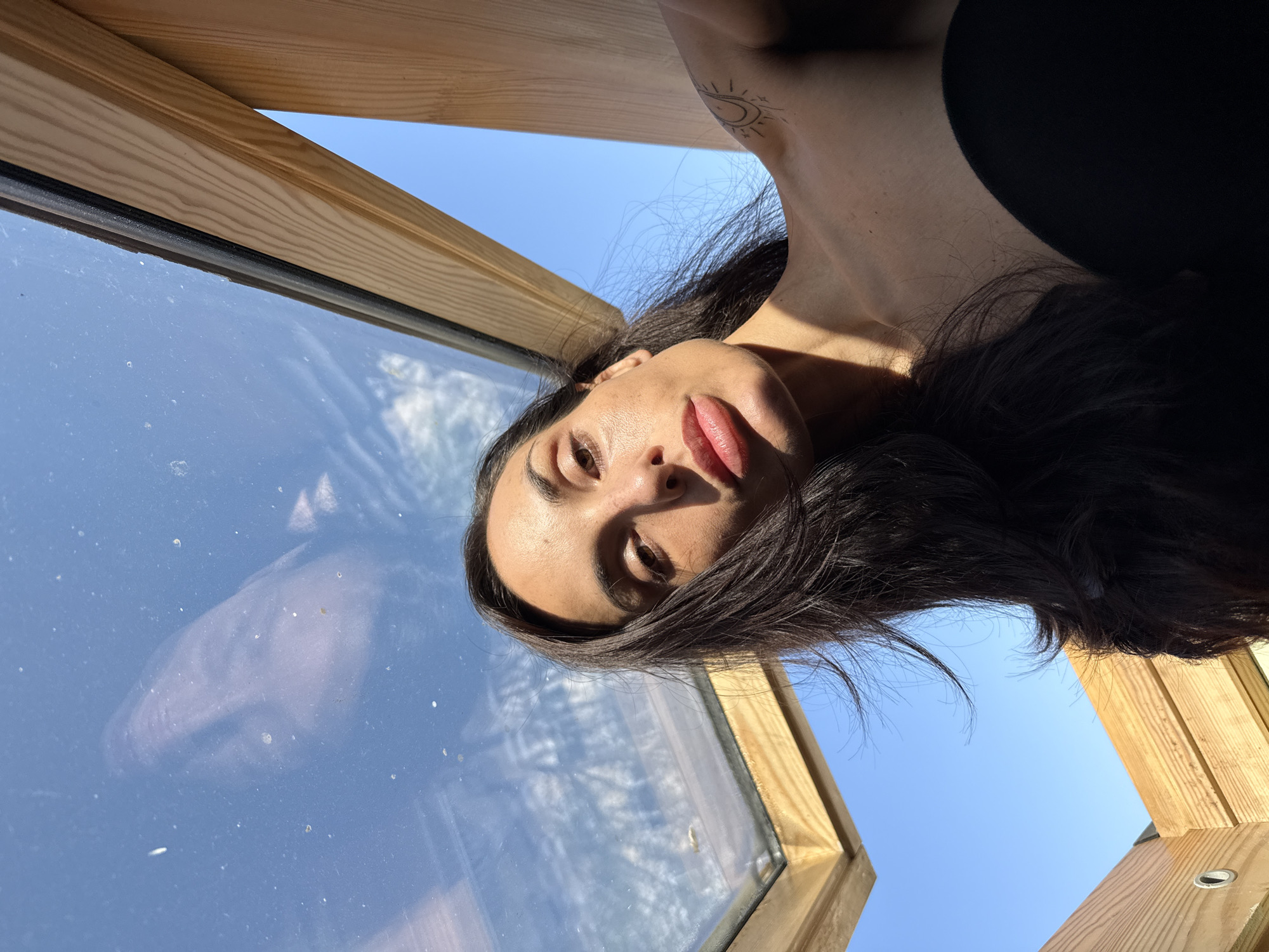 A close-up, upward-angled shot features a young woman looking directly into the camera, her head inverted relative to the clear blue sky visible through a wooden-framed window or skylight. Her dark hair frames her face, which is bathed in bright sunlight, accentuating her dark eyes and full, glossy lips. A delicate tattoo depicting an intertwined sun and crescent moon design is visible on her upper right chest/shoulder.
The scene is set on a bright, sunny day, indicated by the brilliant blue sky and the strong highlights and shadows cast on her skin. The wooden frame suggests an interior space looking out, possibly a room in Pleven, Bulgaria, with a skylight or an unusually positioned window. A dusty glass pane reflects a faint, distorted profile of the woman's face. The overall composition creates a dynamic and intimate portrait, capturing a moment of natural light and serene contemplation.
