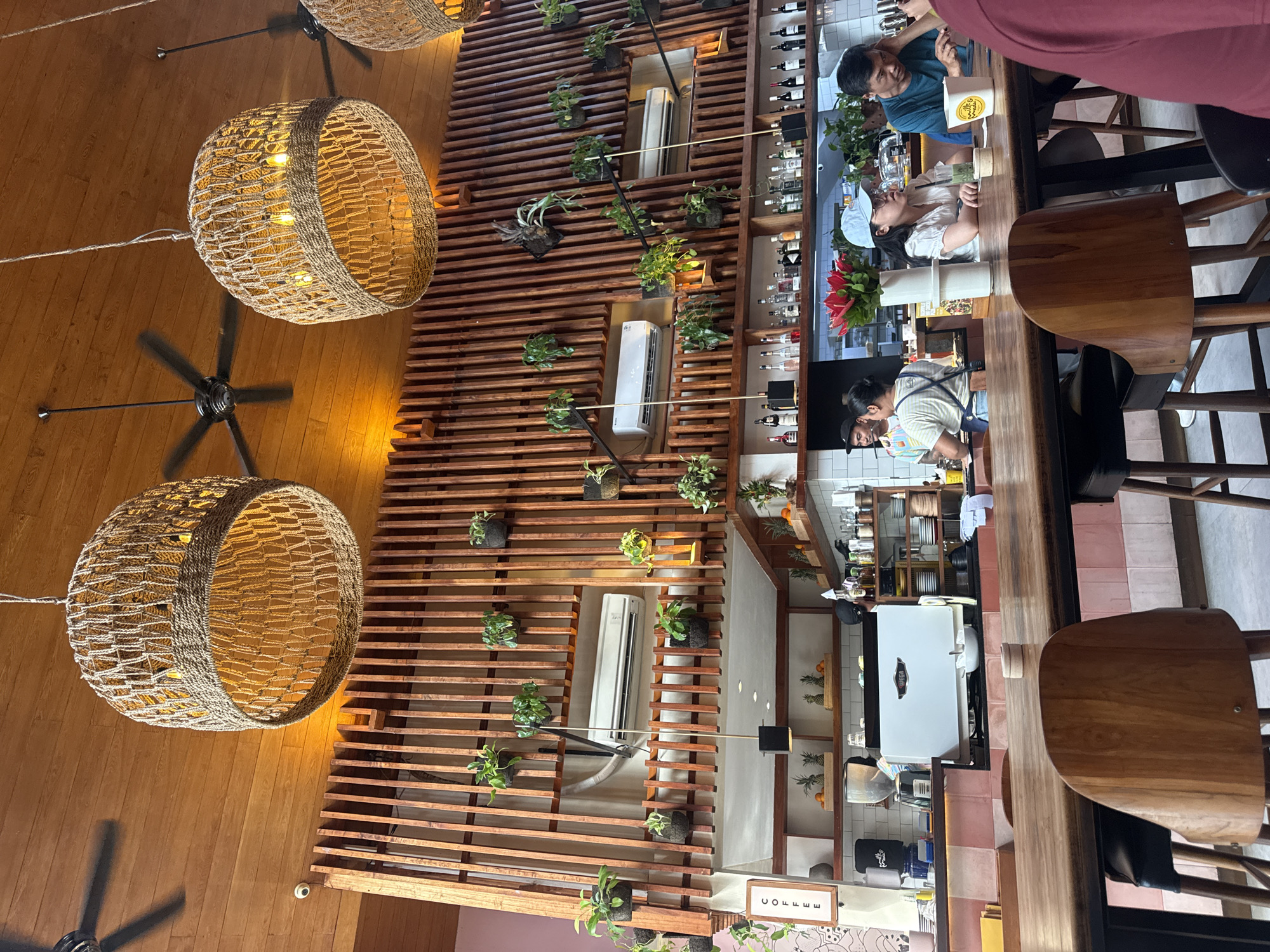 The image shows the interior of a restaurant located in the Ubud District of Indonesia. The scene is shot from below, angled upwards towards the ceiling. Dominating the upper left are two large, woven, basket-like light fixtures hanging from the ceiling. Between them is a black ceiling fan. The ceiling and walls are made of wood.
Running vertically through the middle is a decorative wooden slat wall interspersed with potted plants and three white air conditioning units. To the right of the slat wall is a bar area. Several people are seated at the bar, and a barista is working behind the counter. Bottles of various beverages are displayed on shelves behind the bar. The bar stools have wooden seats and black metal legs. Behind the bar area is a view into a kitchen with tiled walls and shelves.
The image suggests a contemporary restaurant interior with a blend of natural and industrial design elements.
