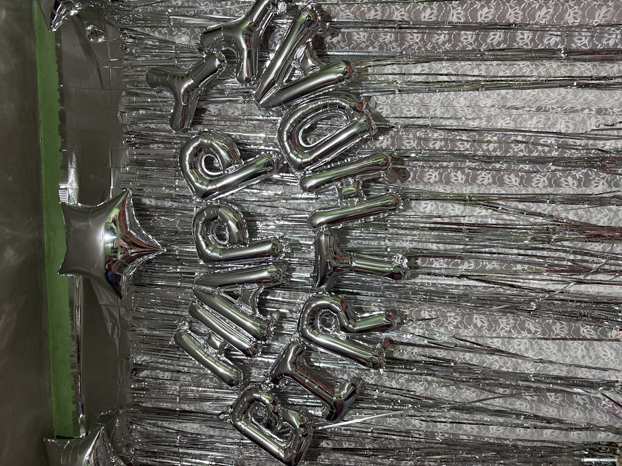 A celebratory backdrop features silver foil balloons spelling out "HAPPY BIRTHDAY" arranged on a background of shiny silver fringe curtains layered over a white lace curtain. Two silver star-shaped balloons are visible at the top. The scene is lit, casting reflections on the metallic surfaces. The location is not identifiable as Patlikuhal, India, as there are no discernible geographical markers in the image.