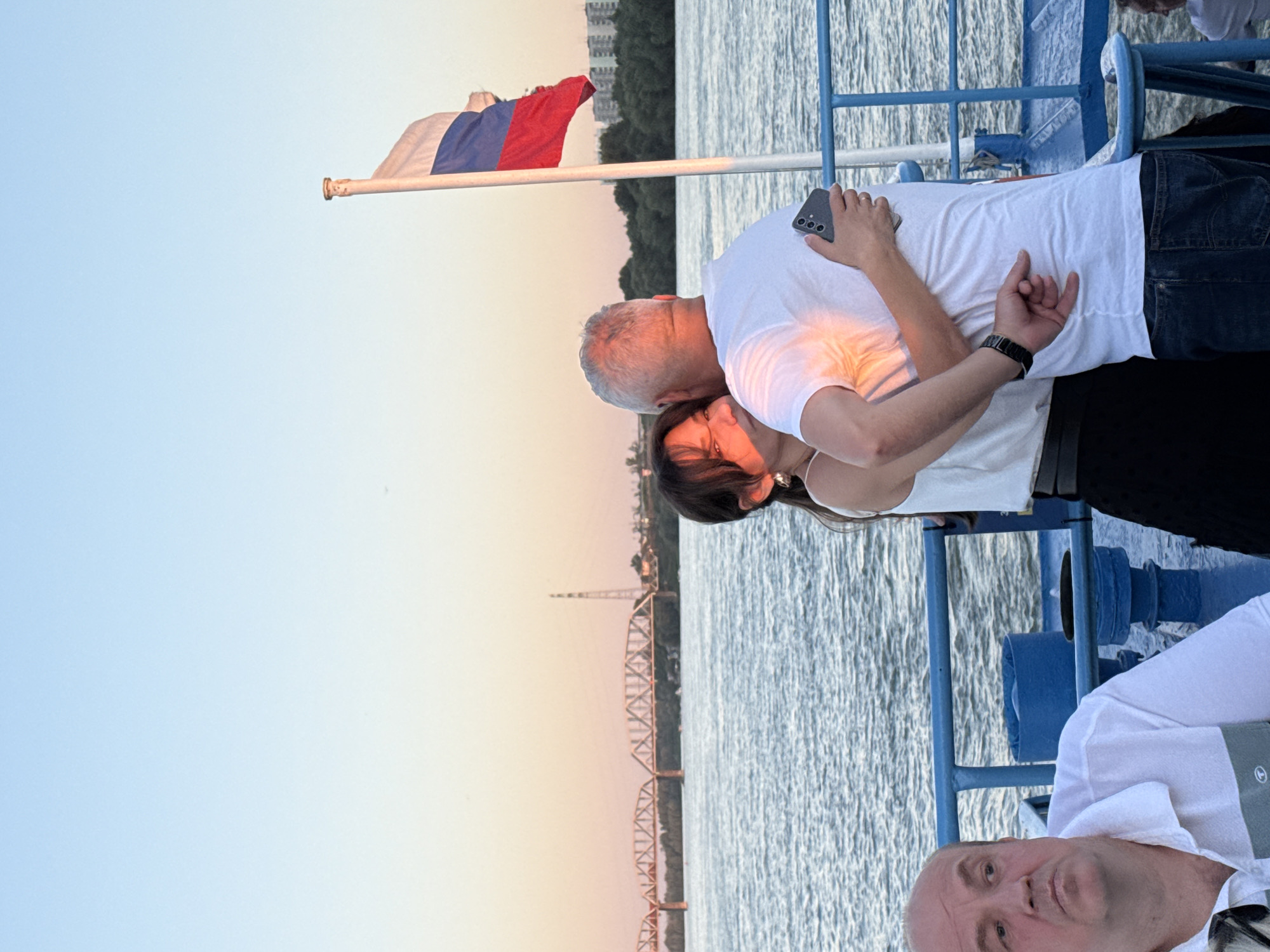 In Novosibirsk, Russia, on a boat, a couple embraces by the Ob River at sunset. The man, with gray hair and a white t-shirt, is holding the woman, who is wearing a white top. The Russian flag flies behind them on a flagpole. In the foreground, a man in a white shirt sits with a serious expression, looking to the left. In the background, a bridge spans the river, and trees line the shore, with buildings visible in the distance. The sky is a pale orange and blue, indicating the end of the day.