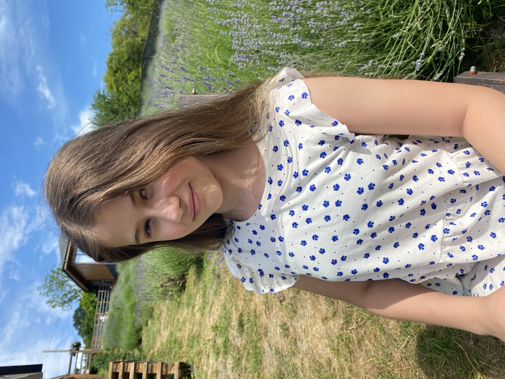 In this media file, a young girl with long, light brown hair is pictured outdoors, likely on a sunny day, given the bright lighting and blue sky with scattered clouds. She is wearing a white dress adorned with a pattern of small blue flowers. Her gaze is directed towards the camera, and she has a subtle smile.
The background suggests a rural or garden setting. Rows of lavender plants are visible, indicating a field or farm. Further back, a wooden structure, possibly a building or shed, is present, along with some trees. The ground is covered with green grass and some dried vegetation, suggesting a warm season. The overall mood is relaxed and natural. The image's orientation is such that the girl and the scene are sideways.