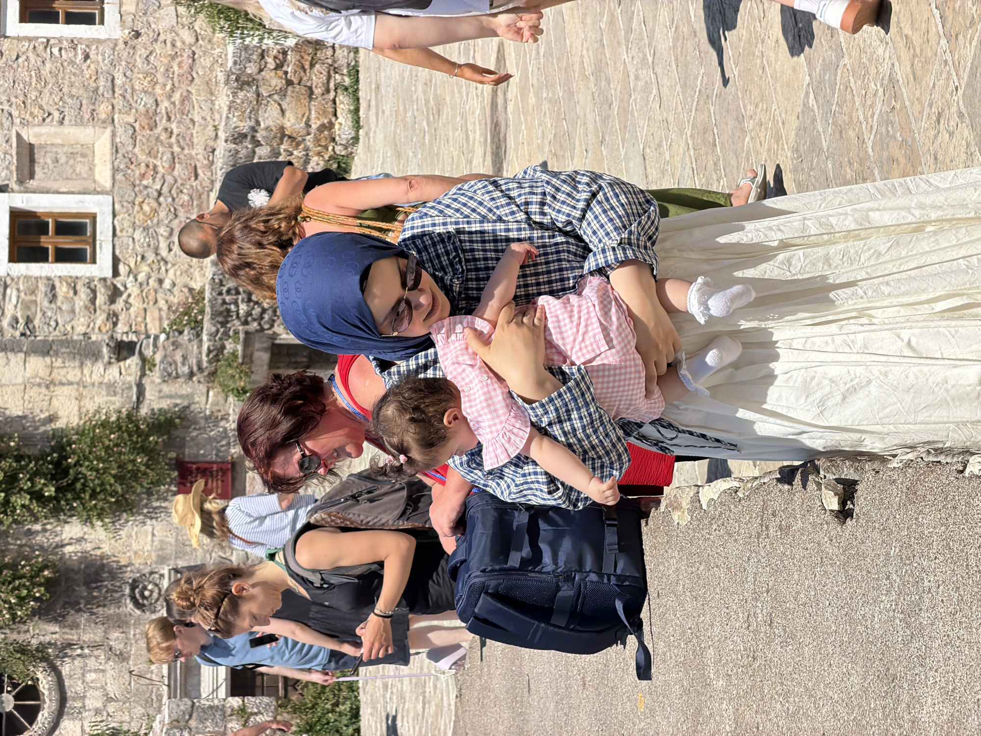 The image captures an outdoor scene in what appears to be a historic, sun-drenched town, likely Budva, Montenegro, given the context. The main subjects are a woman wearing a blue hijab, sunglasses, a blue and white checkered shirt, and a long white skirt, who is holding a baby. The baby, dressed in a pink and white checkered outfit and white ruffled socks, is nestled in her arms, and the woman is smiling down at the child.
Around them, several other individuals are visible. To the left, a woman with auburn hair and sunglasses, wearing a red top, is looking towards the main subjects. A dark blue backpack rests on the ground nearby. Further left and in the foreground, a group of people are walking or standing, including a woman in a black top and a man in a blue shirt. In the background, on the right, other people are present, including a man with a shaved head and sunglasses in a black t-shirt, and another person whose lower legs and feet are visible.
The setting consists of old stone buildings with small wooden-framed windows, typical of ancient or historic architecture. Stone-paved ground forms the walkway. Patches of green foliage grow on the stone walls. The activity suggests a casual outing, possibly sightseeing or a leisurely walk, with families or groups of friends enjoying the sunny weather.
Notable details include the bright, direct sunlight, evidenced by strong shadows cast by people and objects, indicating it's daytime, likely mid-morning or afternoon. The weather appears clear and pleasant. There is a small, unidentifiable red sign mounted on a stone wall in the background, but no legible text is visible in the image. The overall mood seems relaxed and pleasant.