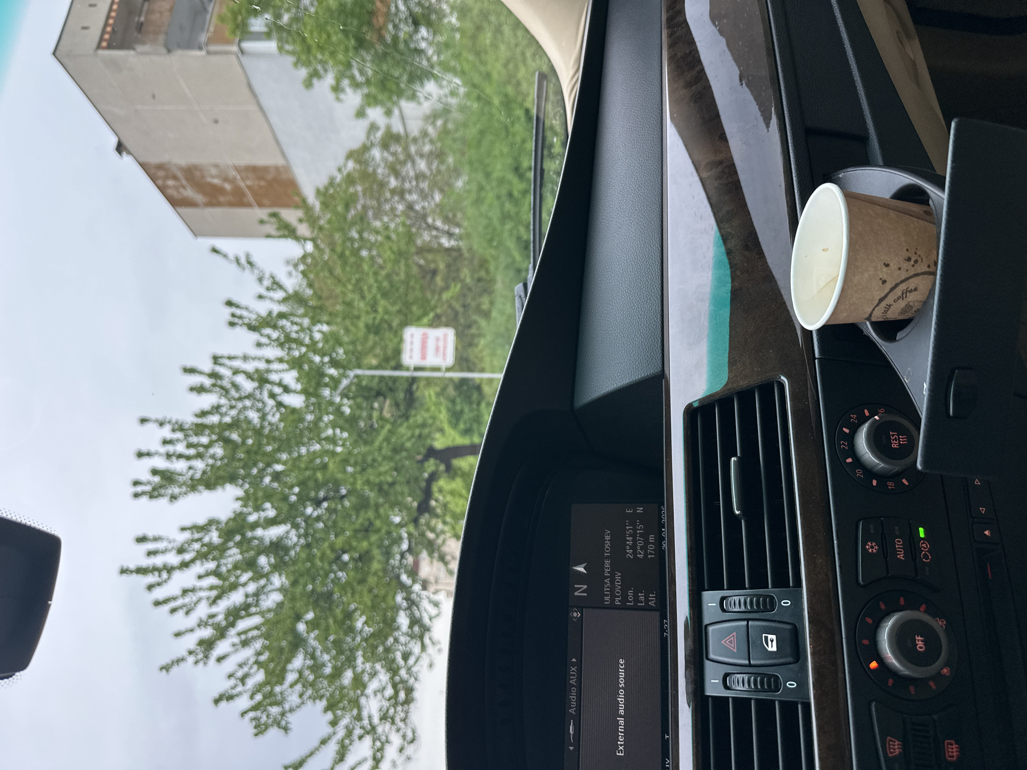 The image provides an interior view from a car, looking out through the windshield and across the dashboard. The perspective is from the front passenger side, showing a detailed view of the car's right-hand side dashboard, including air vents, climate controls, and an infotainment screen. A brown paper cup, with "talk coffee" partially visible, is placed in a cup holder.
The car's infotainment screen is active, displaying navigation information. Visible text on the screen reads "ULITSA PERE TOSHEV", "PLOVDIV", "Lon. 24°44'51" E", "Lat. 42°07'15" N", and "Alt. 170 m", confirming the location as Plovdiv, Bulgaria, specifically on Pere Toshev Street. The screen also shows the time "7:27" and a partial date "30.04.2026", along with "Audio AUX" and "External audio source". The climate control panel shows "AUTO" engaged, temperature settings from "16" to "24" degrees, "REST", and "OFF" buttons.
Beyond the windshield, the scene depicts an urban or suburban area under an overcast sky. Lush green trees, some out of focus, line what appears to be a street. In the background, a multi-story building with a light-colored facade is visible. A white sign with red text, largely illegible, stands amidst the trees. A windshield wiper is visible on the driver's side. The overall impression is of a car parked or stopped during a cloudy day in Plovdiv.