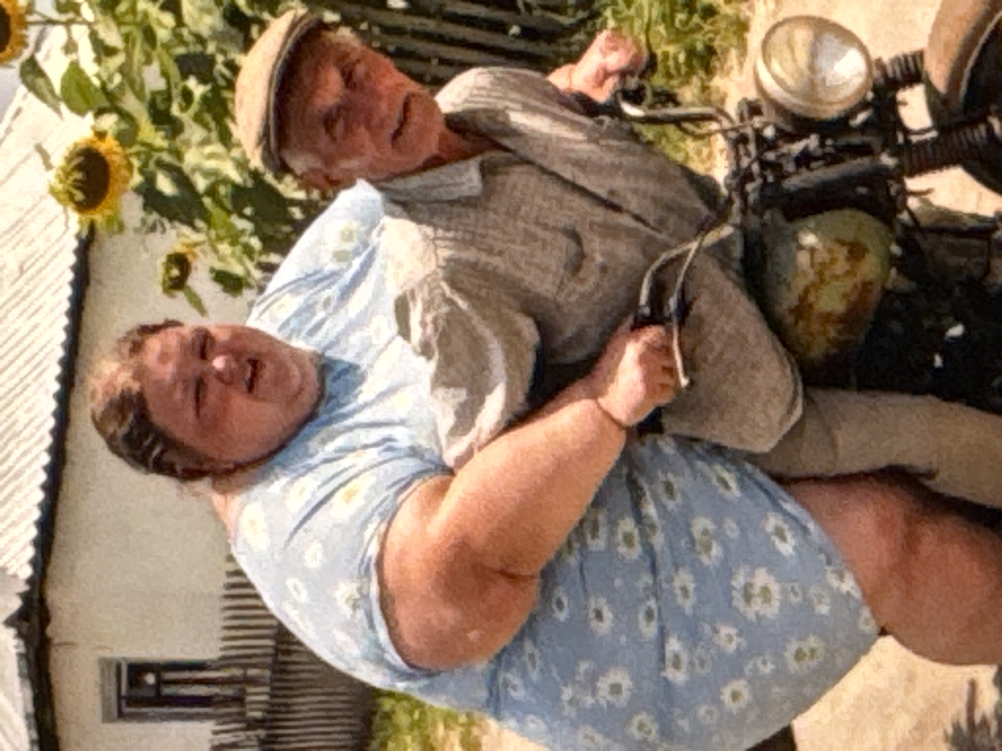 An elderly man wearing a cap and a patterned button-down shirt operates a vintage motorcycle. A woman, significantly larger than the man, is seated behind him on the motorcycle. She is wearing a light blue floral print dress. The scene appears to be outdoors, with a rustic house and sunflowers visible in the background, suggesting a rural setting.