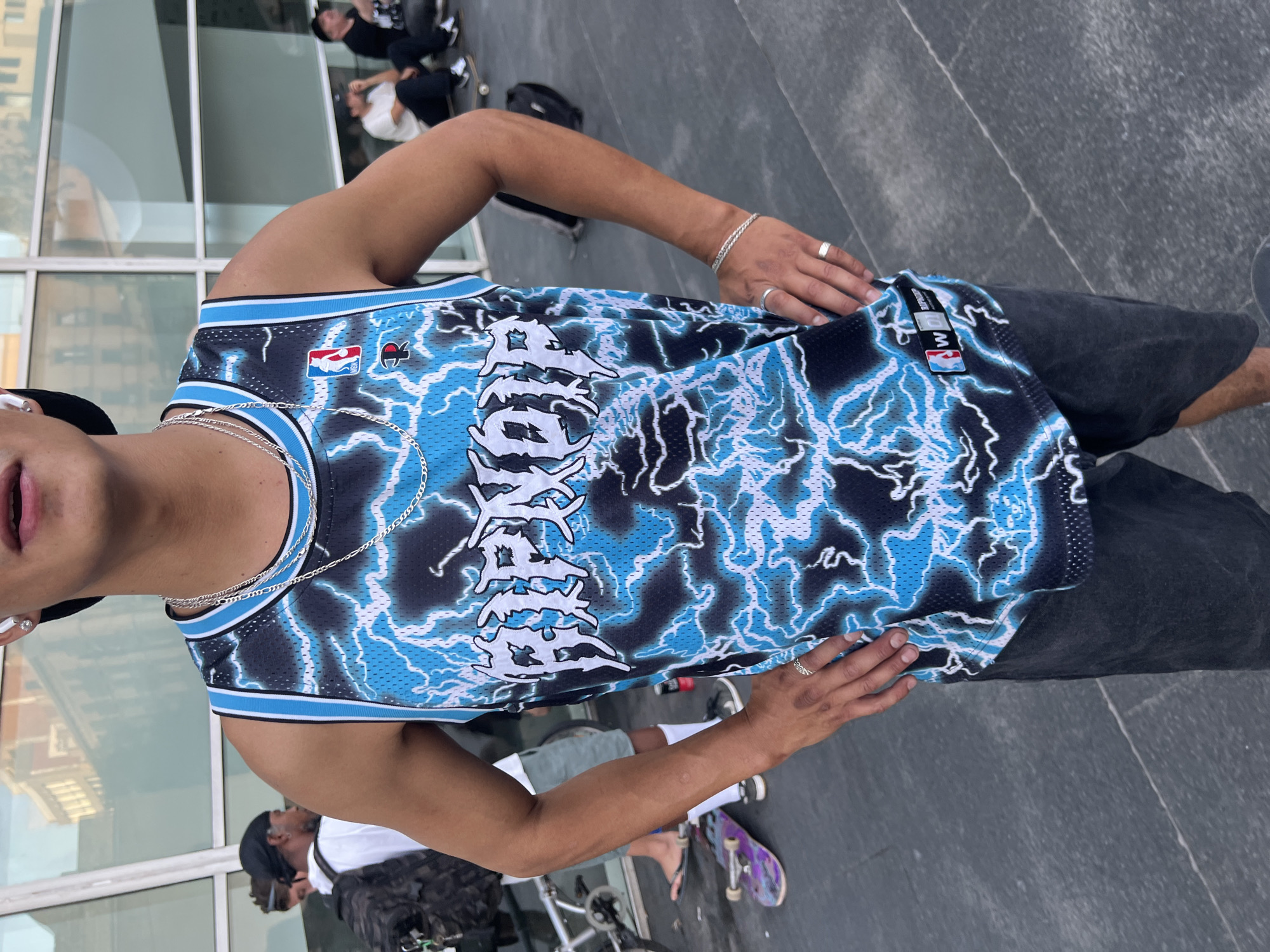 The image shows a person wearing a black and blue lightning-print basketball jersey with "RIPNDIP" written in white across the chest. They are also wearing dark shorts, silver necklaces, and rings. The person is standing in front of a modern building with large glass windows. In the background, other people are visible, some sitting and others walking. There are also skateboards and bicycles in the scene. The ground appears to be paved with large, dark tiles. The lighting suggests it is daytime, with a bright sky. The scene is likely an urban public space, possibly a plaza or an area outside a commercial building. The presence of skateboards and casual attire hints at a youthful or counter-culture gathering. The architecture and general ambiance could be found in various cities, and without further context, pinpointing a specific location like Barcelona is not possible from the visual information alone.