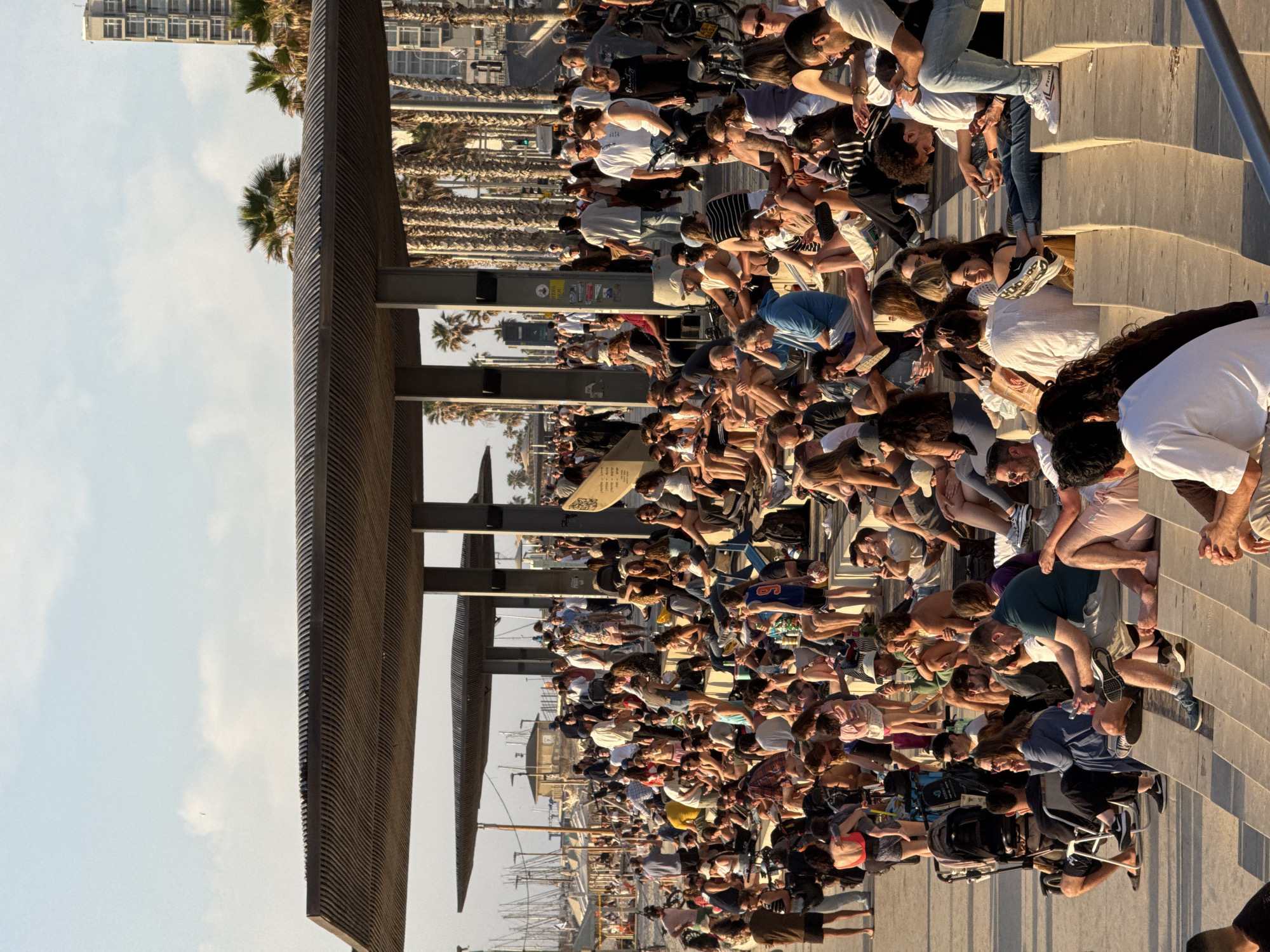 A large crowd of people gathers at the Tel Aviv Marina promenade. They are sitting and standing on stone steps. Many are facing the sea. A slatted, wooden structure provides shade. Palm trees line the promenade. In the background, a high-rise building is visible. The scene is bathed in sunlight. Boats are visible at the marina.