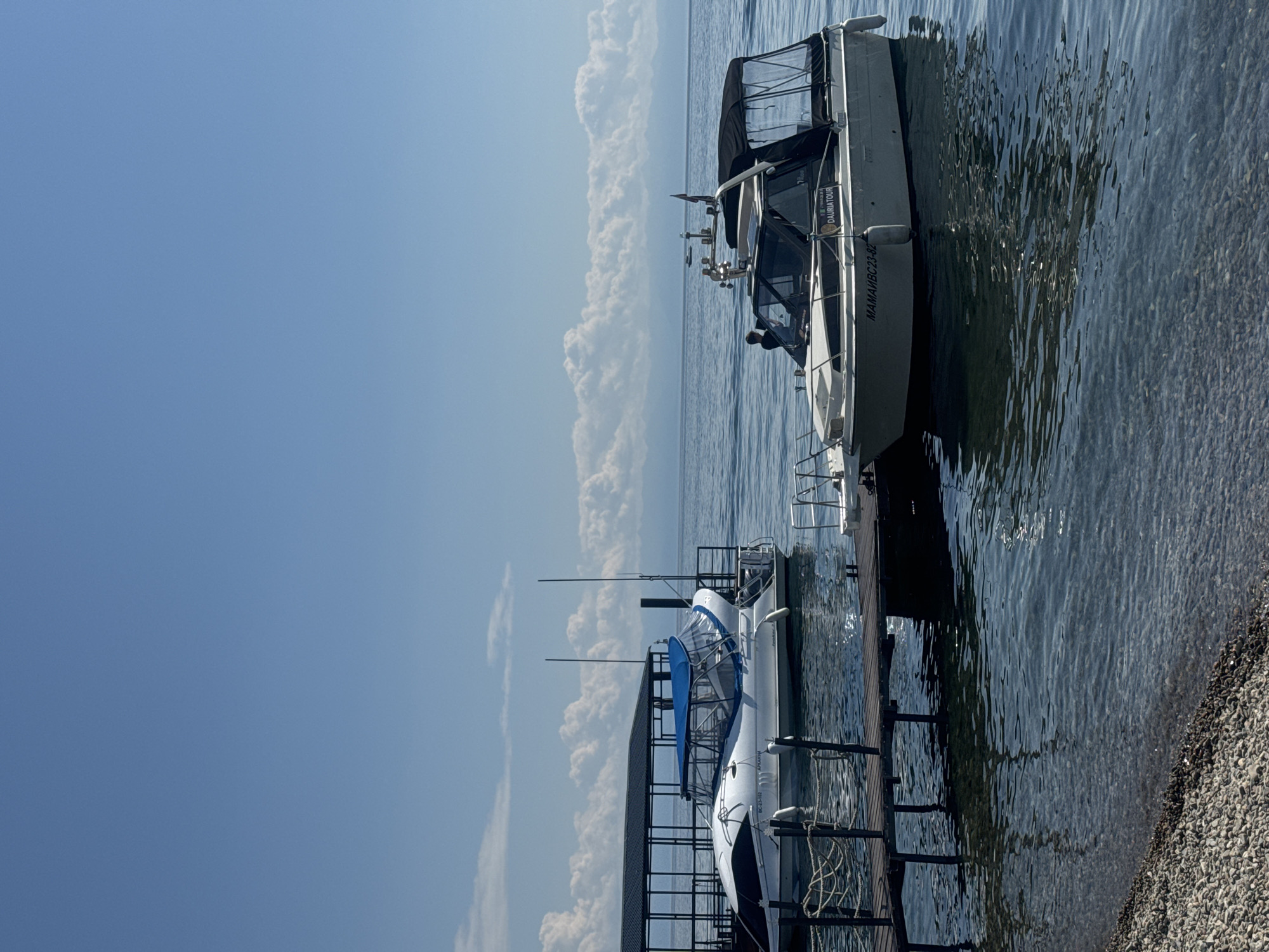 A wide shot captures a serene view of Lake Baikal in Listvyanka, Russia, under a clear blue sky dotted with white, puffy clouds. Two boats are docked. In the foreground, a sleek white and blue motorboat is moored to a wooden pier, its blue canopy providing shade. The boat has a black stripe and an inscription that appears to read "MAMAИBC23-82" and "DAURIATOUR". Behind it, another white motorboat with a blue canopy is also docked, partially obscured by the first boat. The water is calm and clear, with the pebbled shoreline visible in the lower left corner. In the distance, a faint mountain range is visible on the horizon. A person is seated on the deck of the closer boat, appearing to be the pilot.