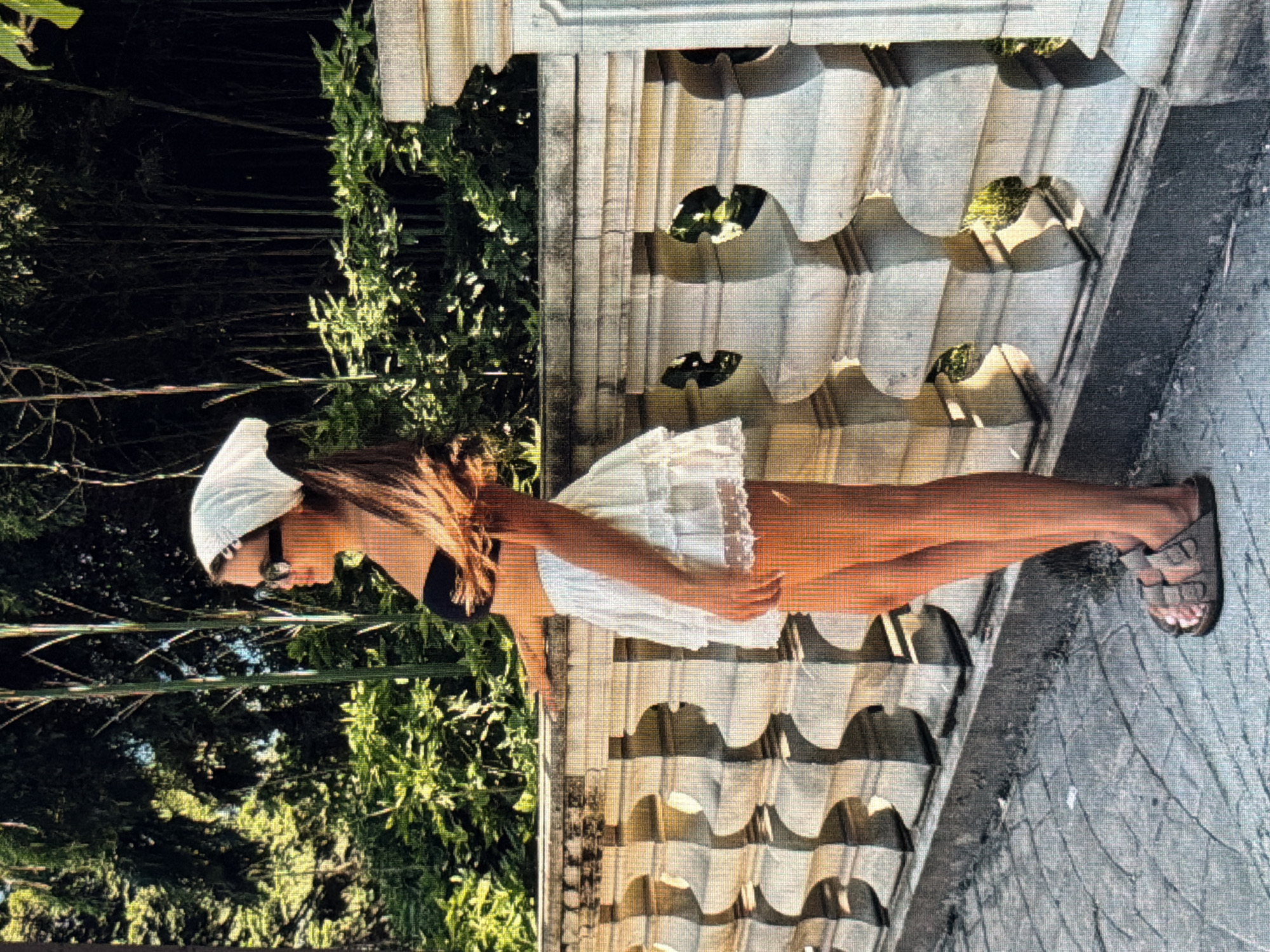 A young woman stands on a paved walkway next to a stone balustrade. She is wearing a white bandana on her head, sunglasses, a black bandeau top, and a white ruffled skirt. Her hair is long and brown, and it is blowing in the wind. She is looking to her left, with her left arm resting on the balustrade. Behind her is a dense foliage of bamboo and other greenery. The balustrade is made of stone and has decorative carvings. The walkway is paved with cobblestones. The scene appears to be in a park or garden, with the sun casting shadows on the ground. The location is identified as Sirius, Russia.