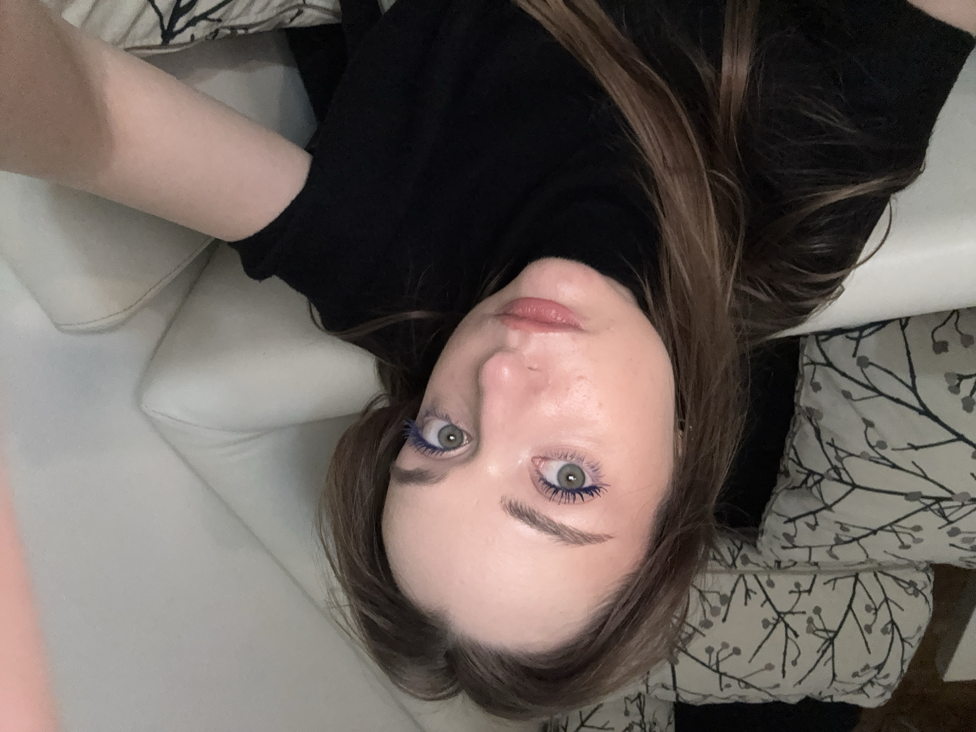 A woman with long brown hair is lying on a white couch. She is wearing a black turtleneck sweater. Her eyes are green and she has blue mascara on her eyelashes. The background includes a patterned pillow. The location is unknown and cannot be determined from the image.