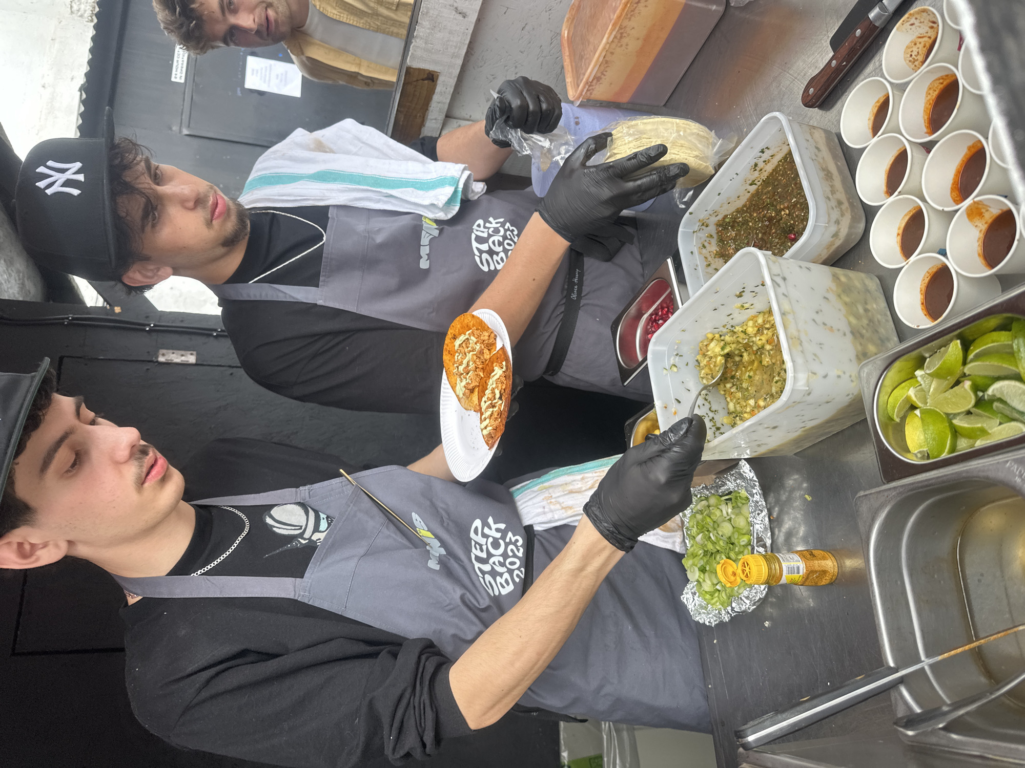 The image shows two men preparing food in a food stall. Both are wearing black long-sleeved shirts and grey aprons. The aprons have white writing on them. They are both wearing black gloves. The man in the foreground is adding ingredients to a foil package containing chopped green onions. To the upper right, the man in the background is preparing tacos. He is holding a bag of tortillas in his left hand. There are several containers with various ingredients, including salsa, lime wedges, and small cups filled with a reddish-brown liquid. The food stall appears to be metal, and there is a stainless steel sink to the right. The location appears to be an outdoor food stall in London, United Kingdom.