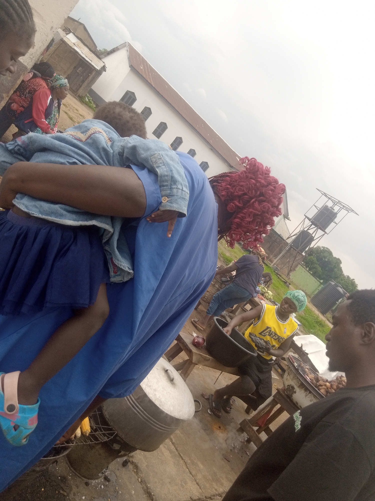 In Barakin Lerawi, Nigeria, the image captures an outdoor scene with several individuals and food preparation activities. A person wearing a blue denim jacket and a blue skirt holds a child. Nearby, another person with bright red, curly hair is visible. In the background, a building with a reddish-brown roof is situated. Several people are engaged in cooking, with large pots and grilling setups present. One individual wears a yellow basketball jersey with "Lakers" text, holding a raw fish. Another person is seen preparing food in a large black basin. A water tower with two tanks stands in the distance, along with other structures and vegetation. The ground appears to be a mixture of dirt and concrete. The lighting suggests it is daytime, with an overcast sky.