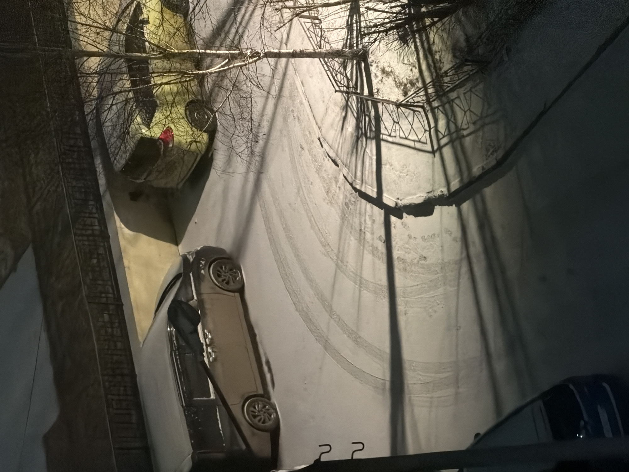 The image shows a snow-covered street scene in Krasnoyarsk, Russia, viewed from an elevated perspective, possibly a balcony or window. Tire tracks are visible in the snow on the street. Two cars are parked along the side of the road. Bare trees and the shadow of a fence or railing are cast across the street. The image is taken at night, with lighting provided by street lamps.