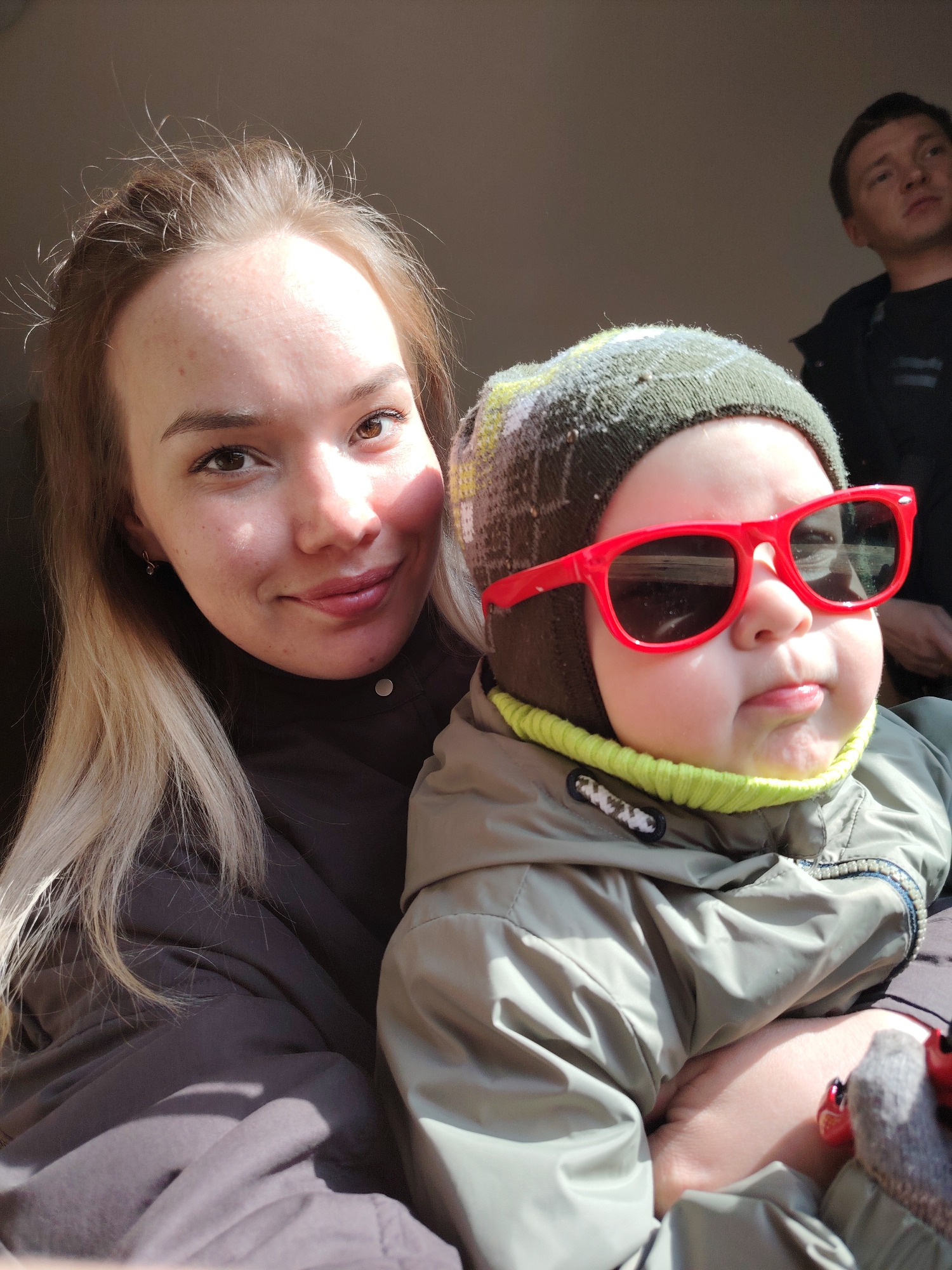 A selfie taken indoors features a woman holding a baby wearing a hat and oversized red sunglasses. The woman has fair skin, brown eyes, and blonde hair styled in a center part. She is wearing a dark jacket. The baby is wearing a green jacket and a patterned hat. In the background, partially visible, is a man with short brown hair and a black jacket. The image is taken indoors and partially shadowed. The picture was taken in the city of Марковский, Russia.
